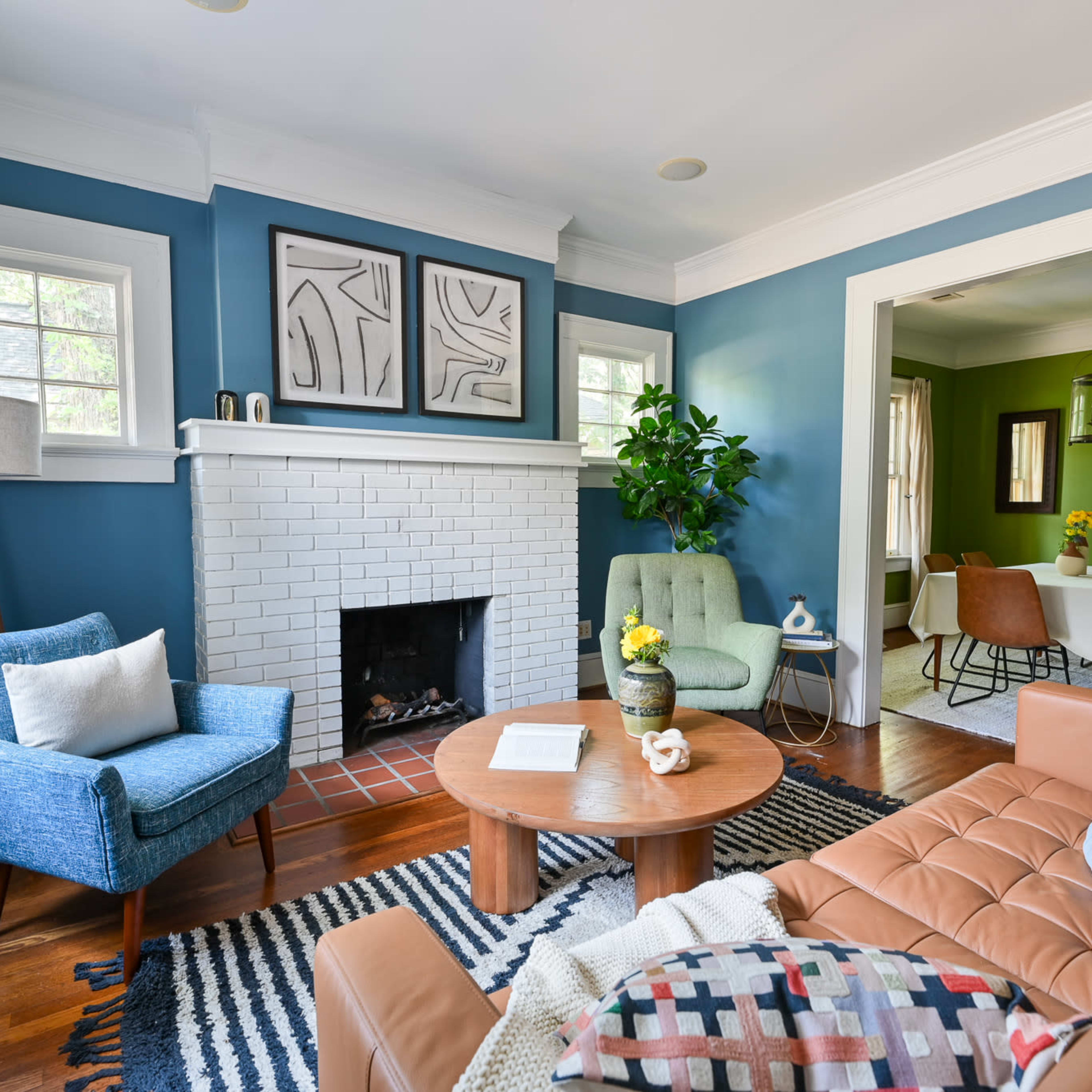 A cozy living room with a brown leather sofa, blue accent chairs, a round coffee table, and a white brick fireplace surrounded by blue walls.