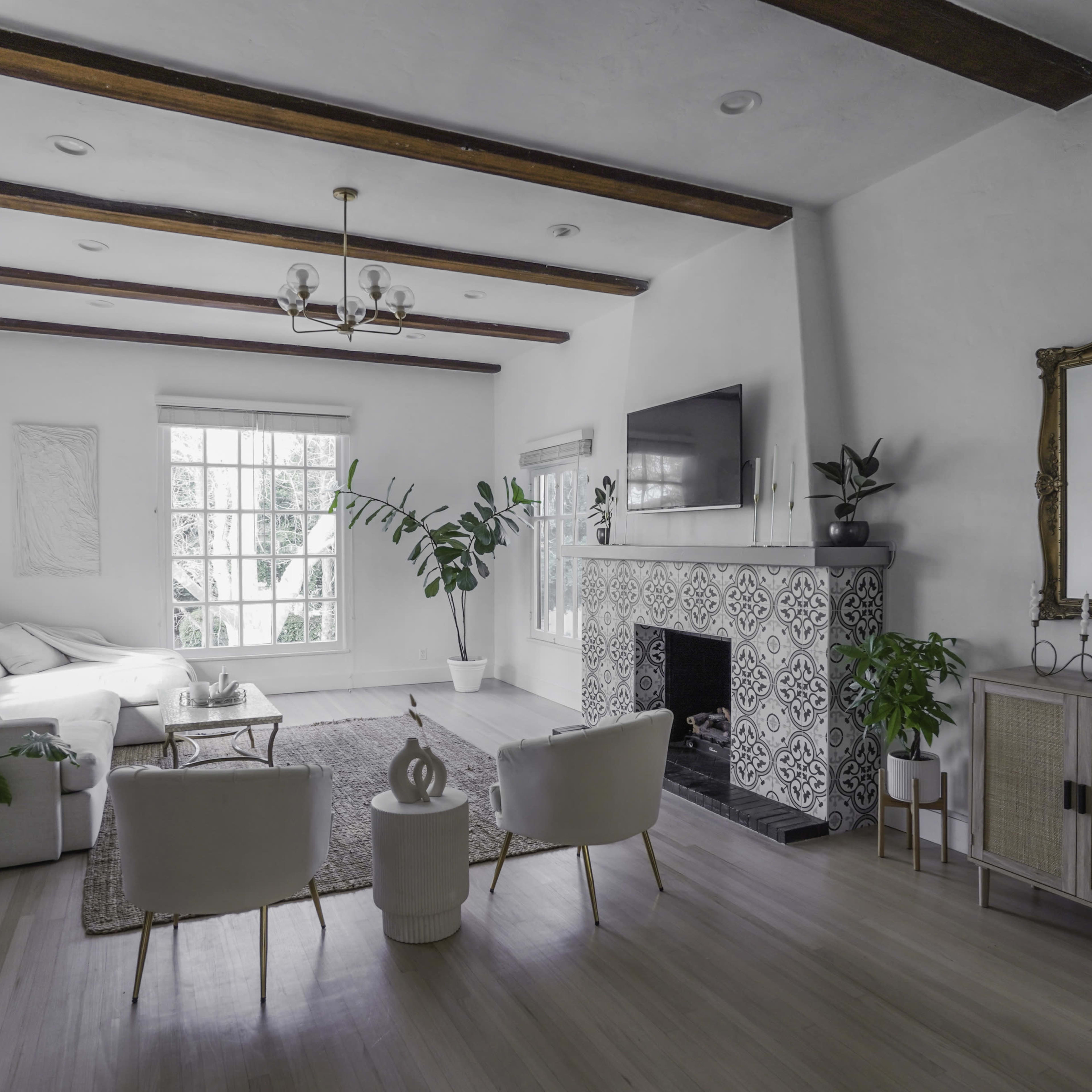 A spacious living room features white walls, wooden beams, modern furniture, and a decorative fireplace.