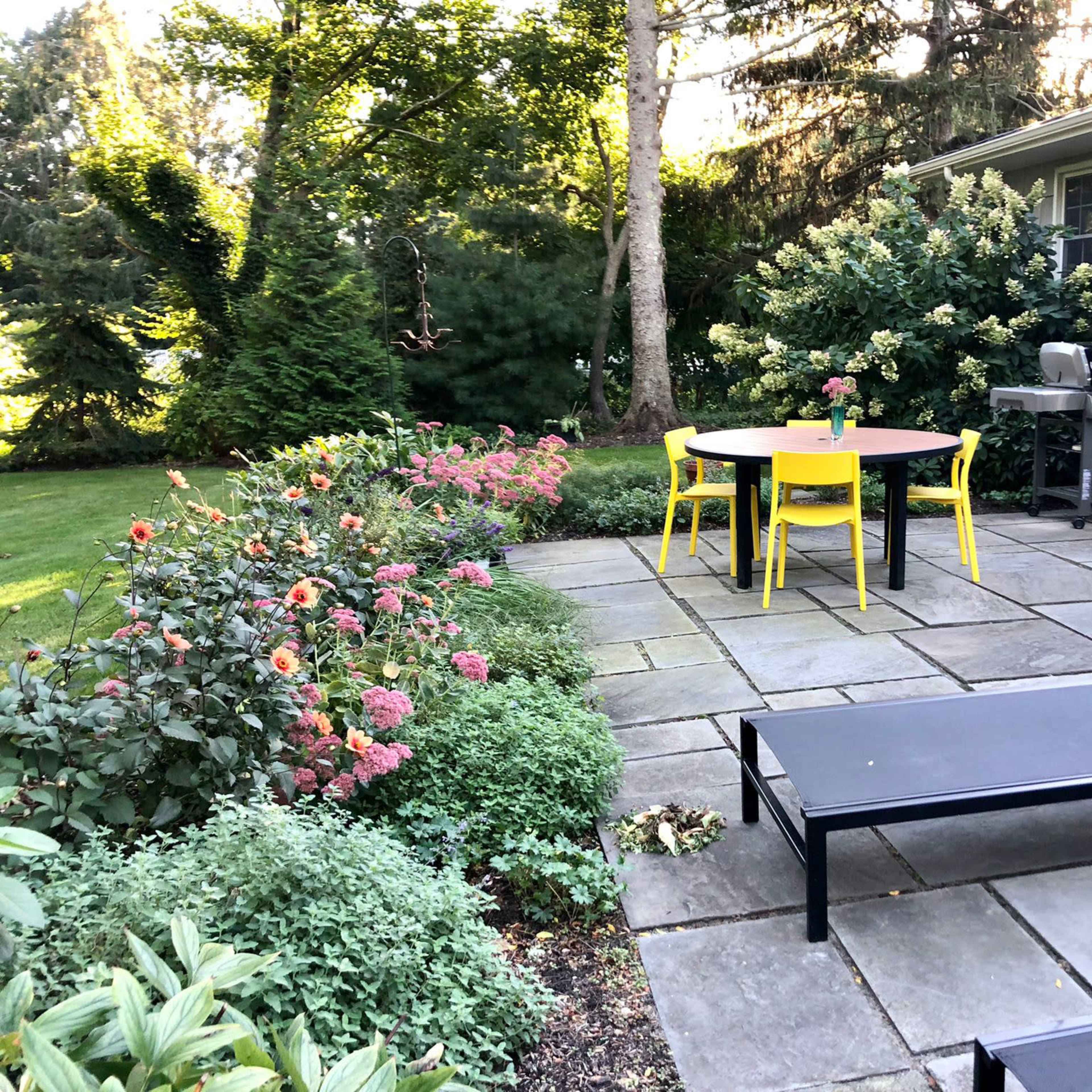 A patio area features a round table with yellow chairs, surrounded by a flower garden and a grill, adjacent to a house.