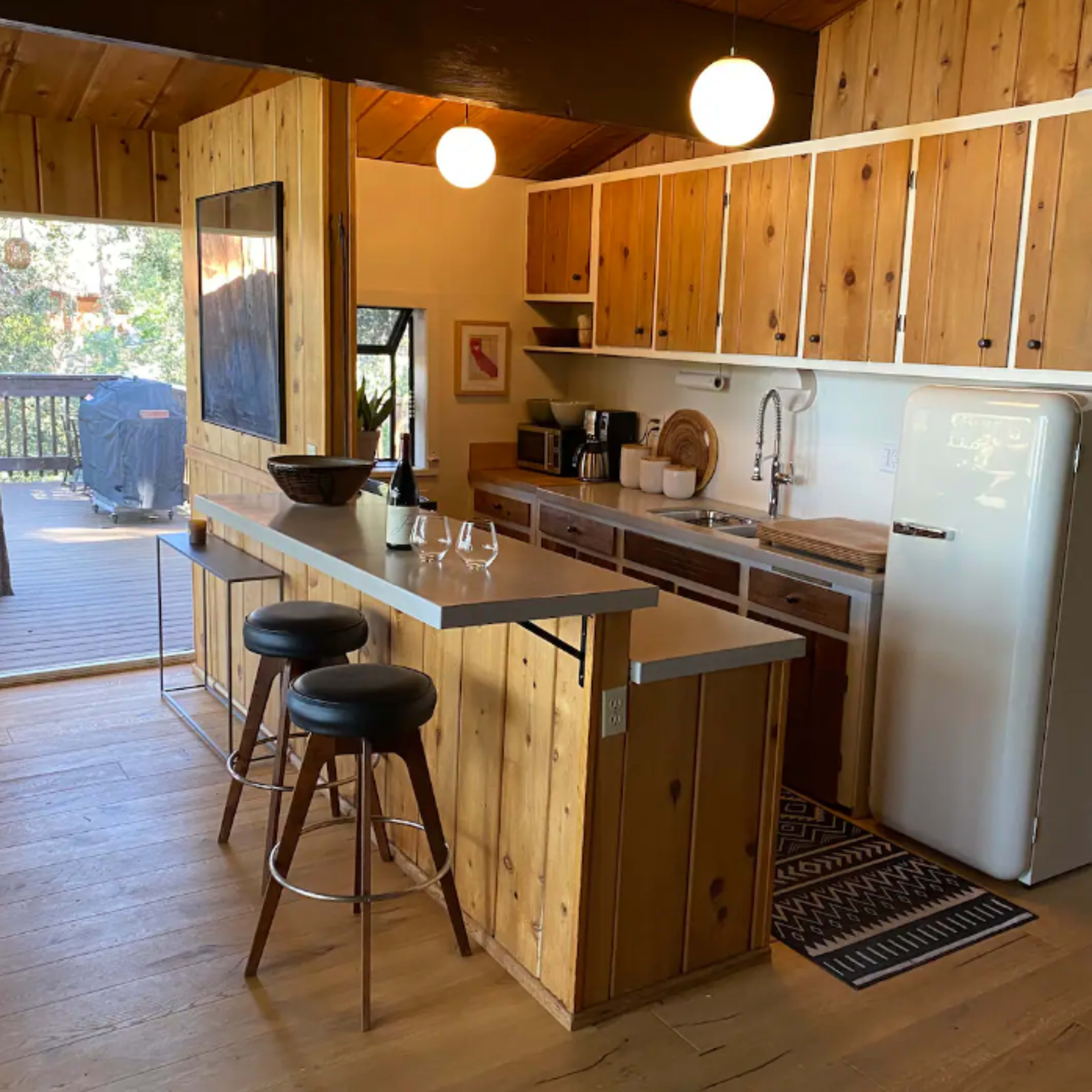 A wooden kitchen with a bar counter and stools, featuring a sink, refrigerator, and cabin-style decor, adjacent to a deck area.