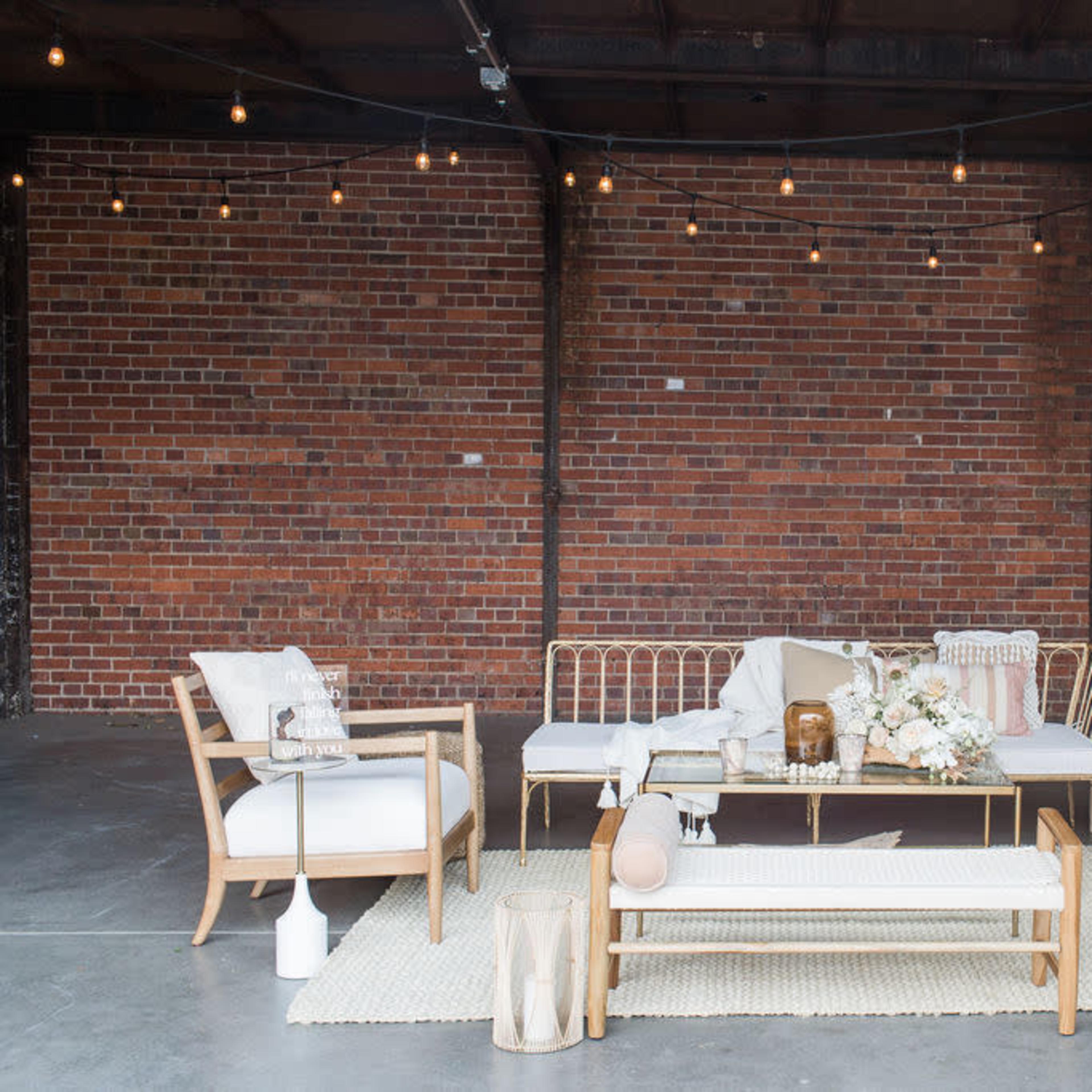 A seating area with wooden furniture, a coffee table, and a floral arrangement is set against a brick wall with string lights overhead.