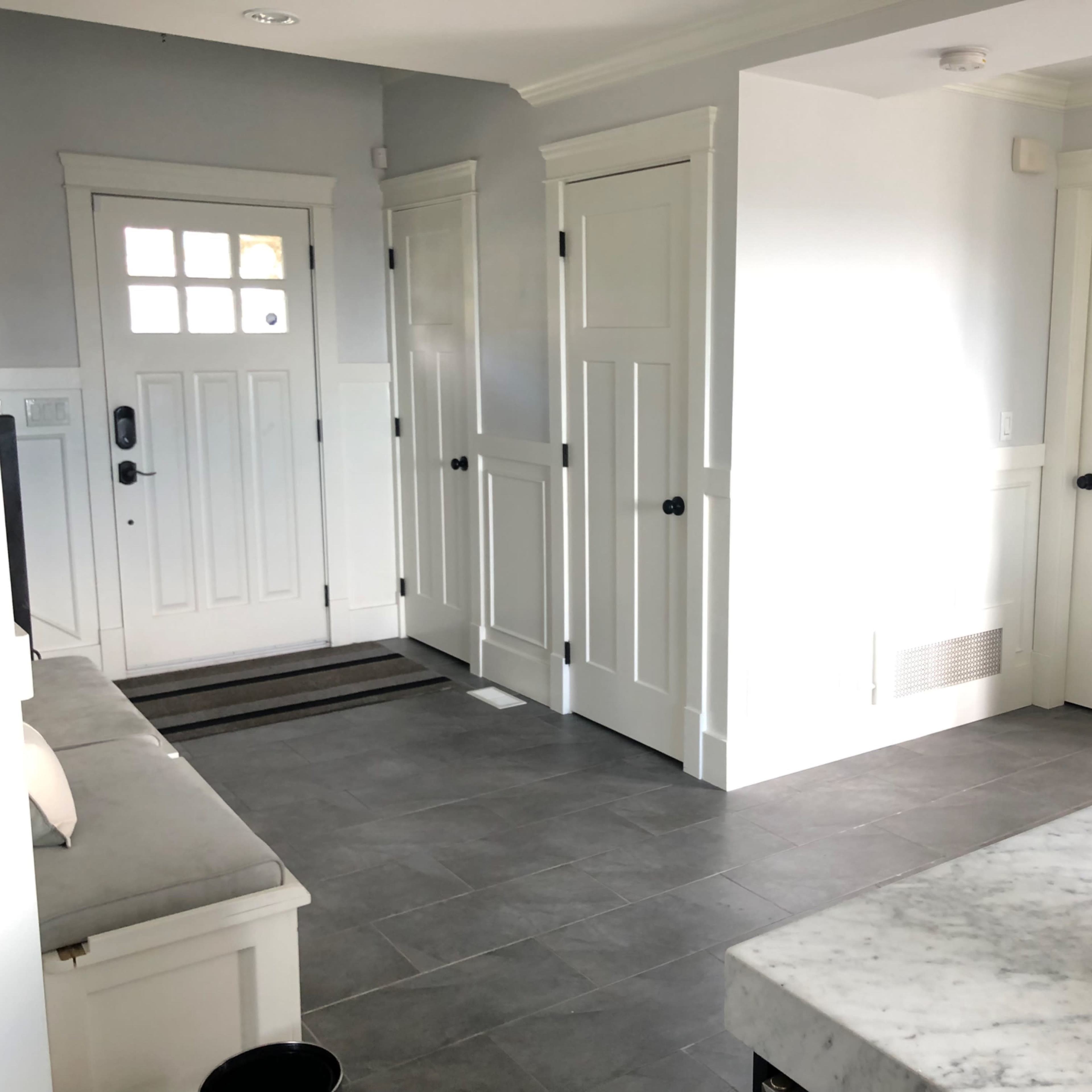 A hallway with multiple closed doors and a front entrance, featuring a gray tiled floor and a white marble countertop in the foreground.
