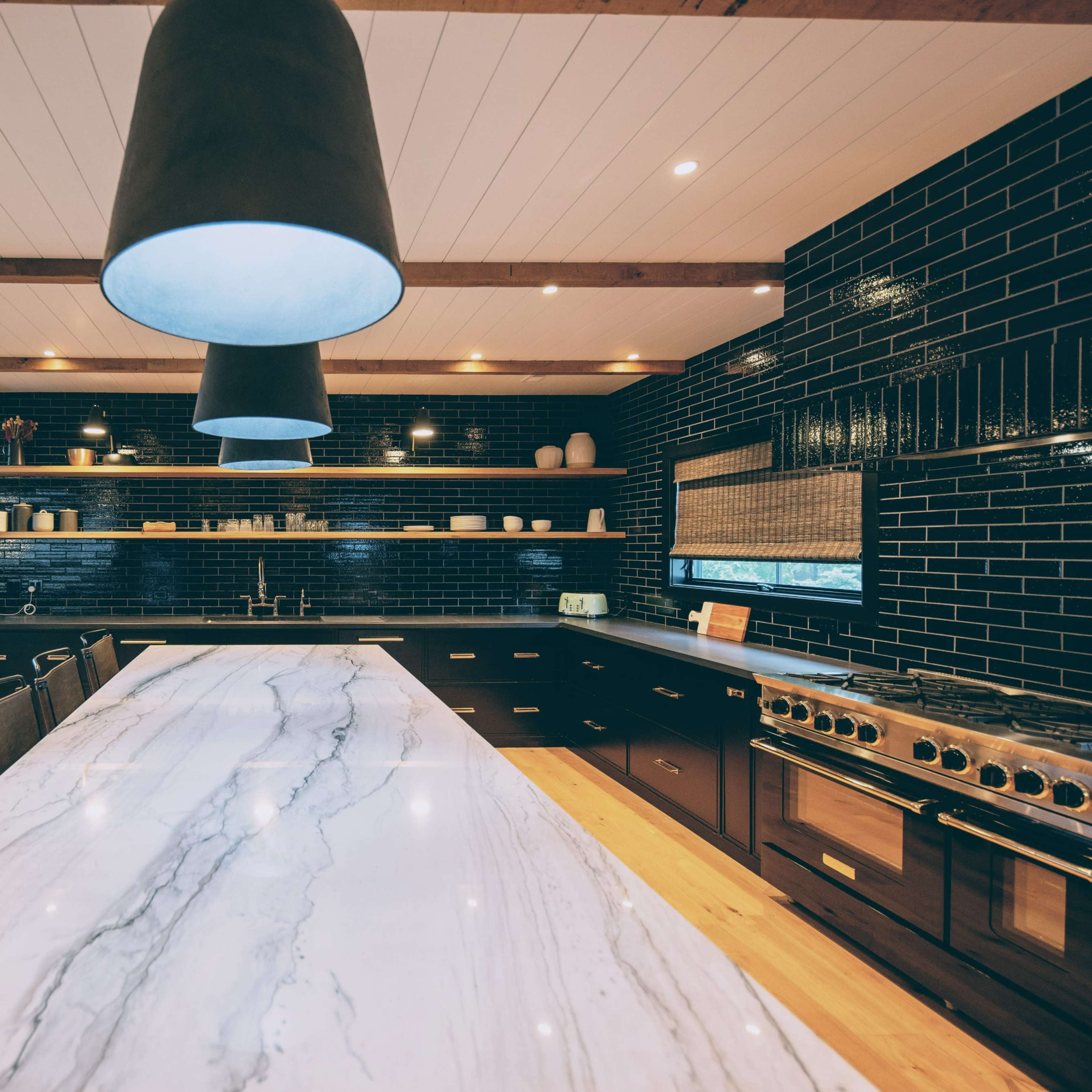 A modern kitchen with a large marble countertop, pendant lights, dark cabinetry, and a staircase in the background.