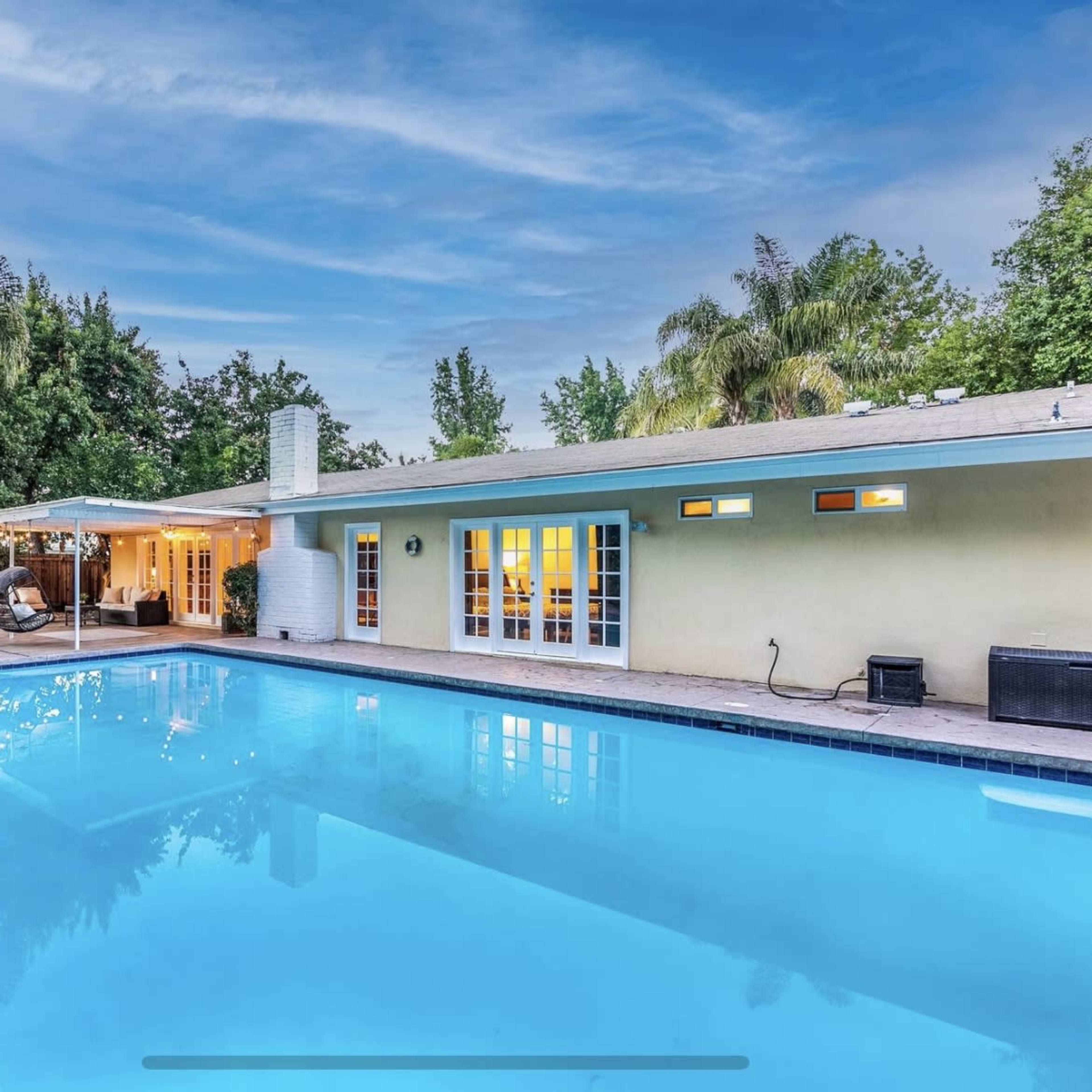 A backyard features a clear pool surrounded by a patio, with a covered seating area and a light-colored house in the background.