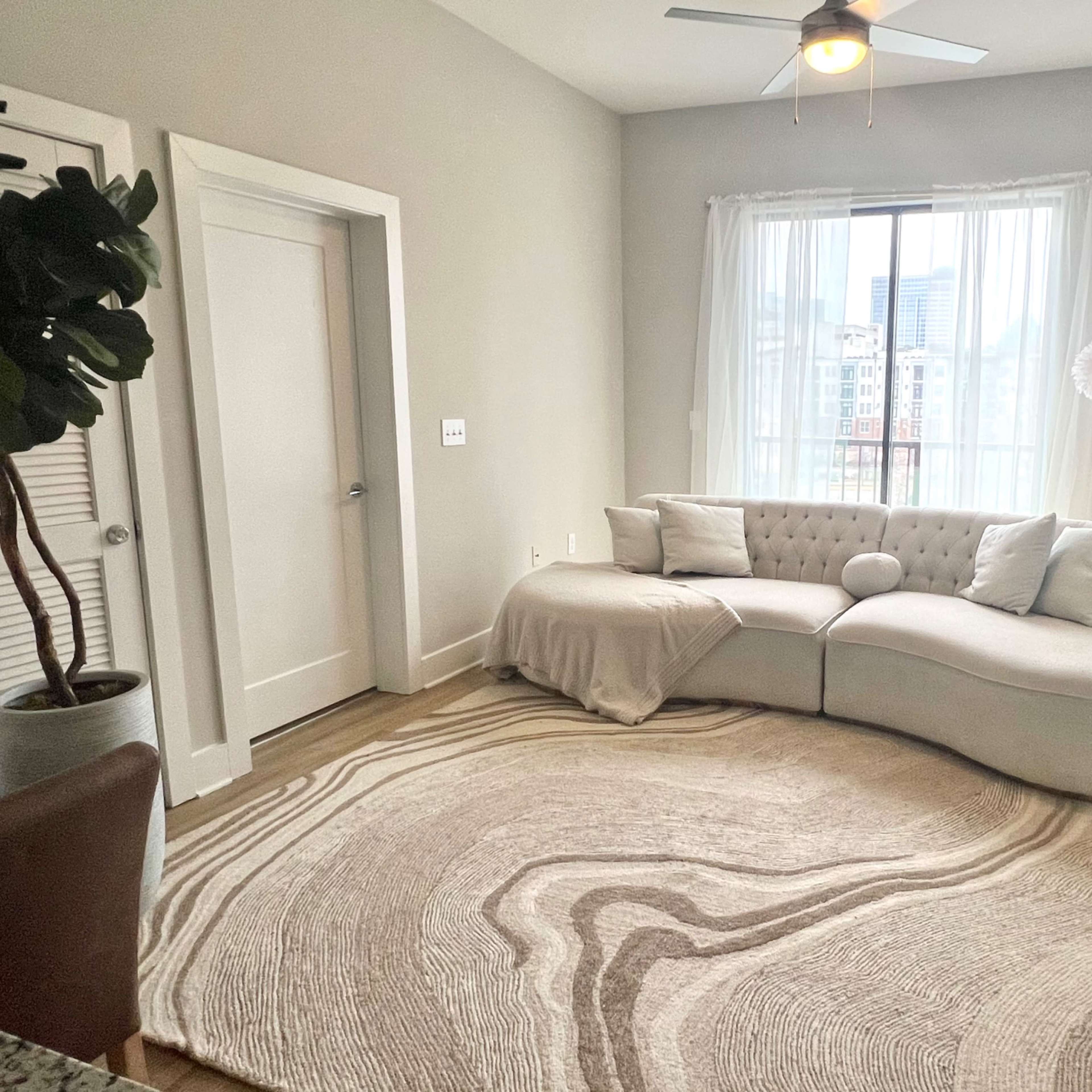 A modern living room featuring a curved white sectional sofa, a round area rug with a wave design, and large windows dressed with sheer curtains.