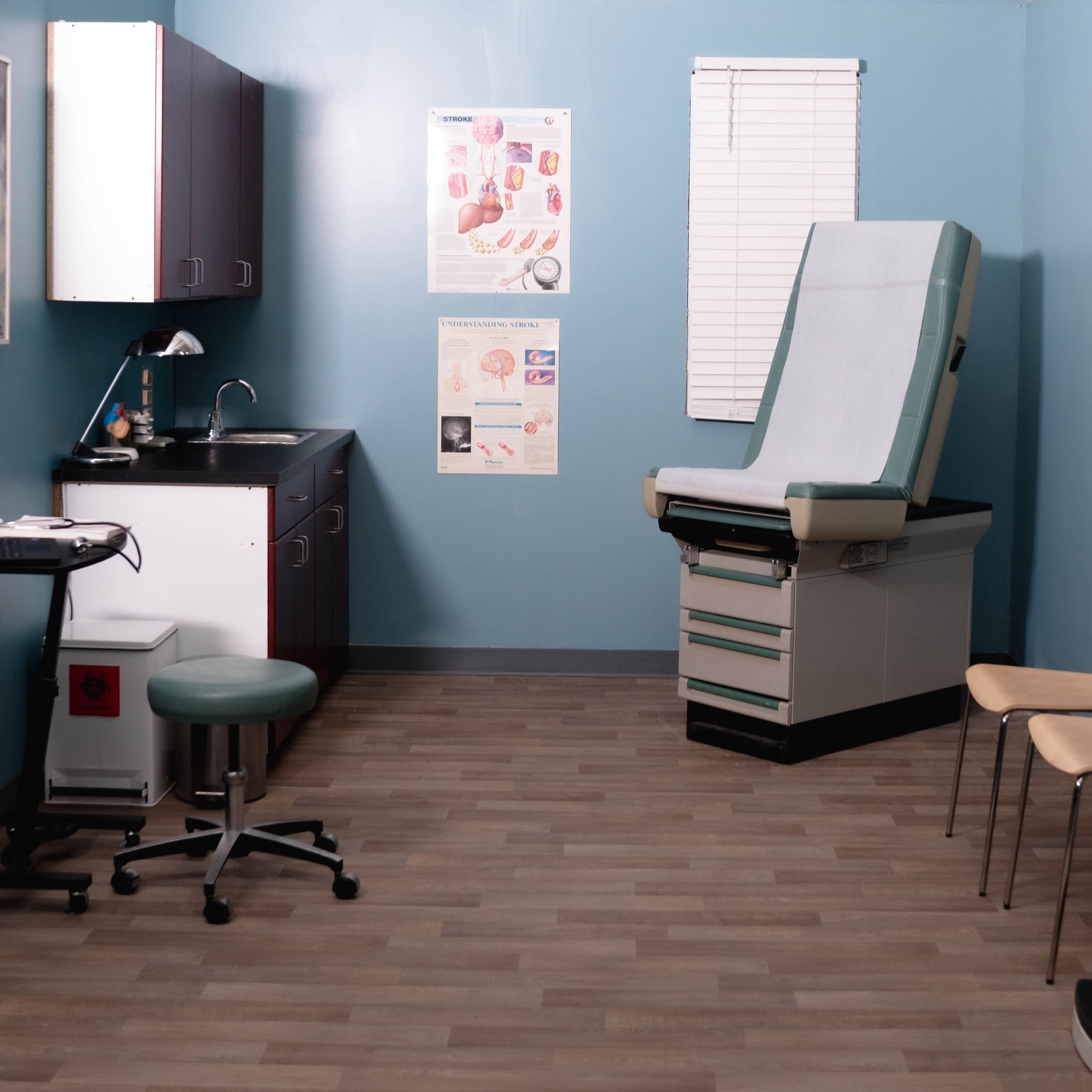 A well-lit medical examination room with an examination table, chairs, a sink, and medical equipment displayed on the walls.