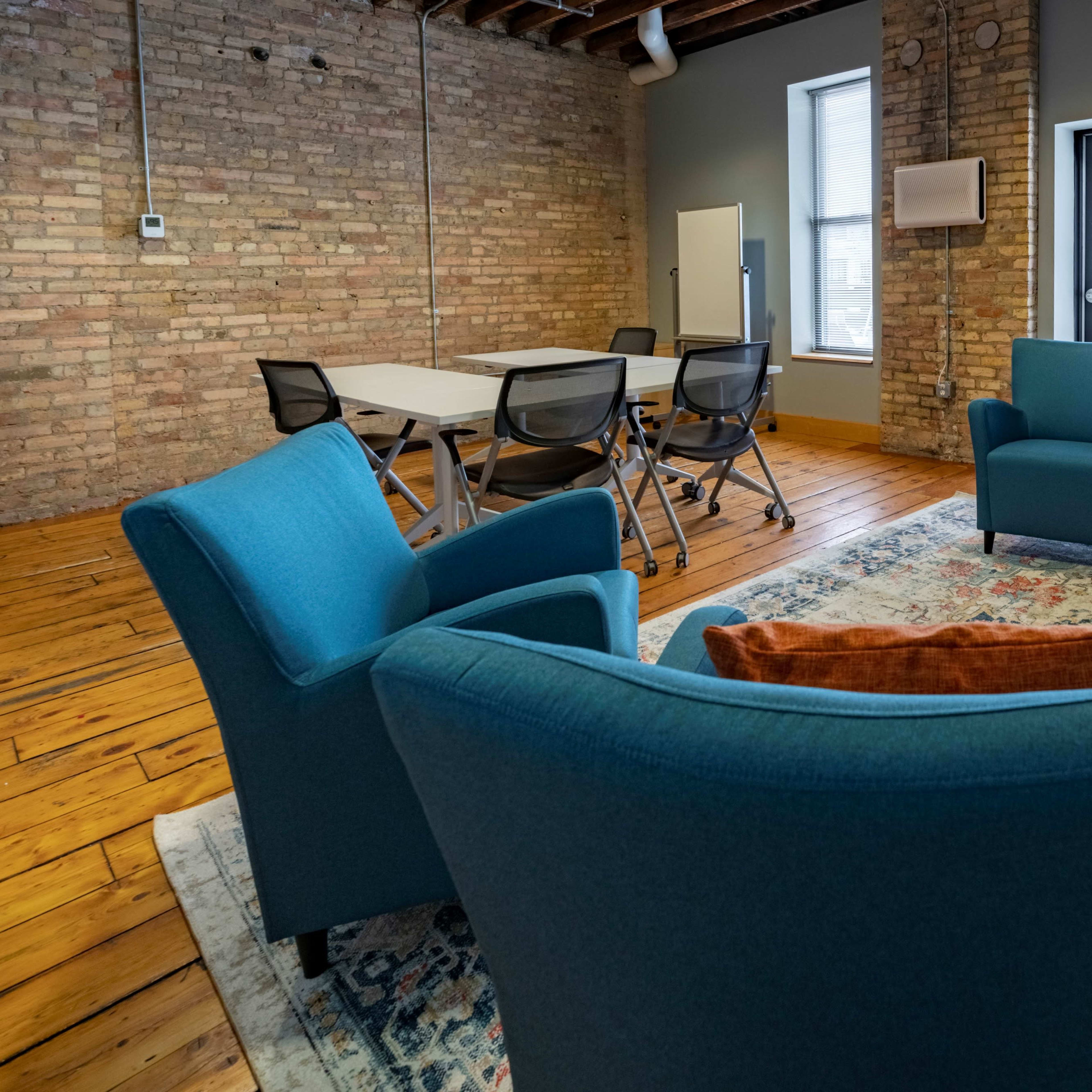 A conference room with four blue chairs arranged around a small table on a patterned rug, with a large table in the background and exposed brick walls.