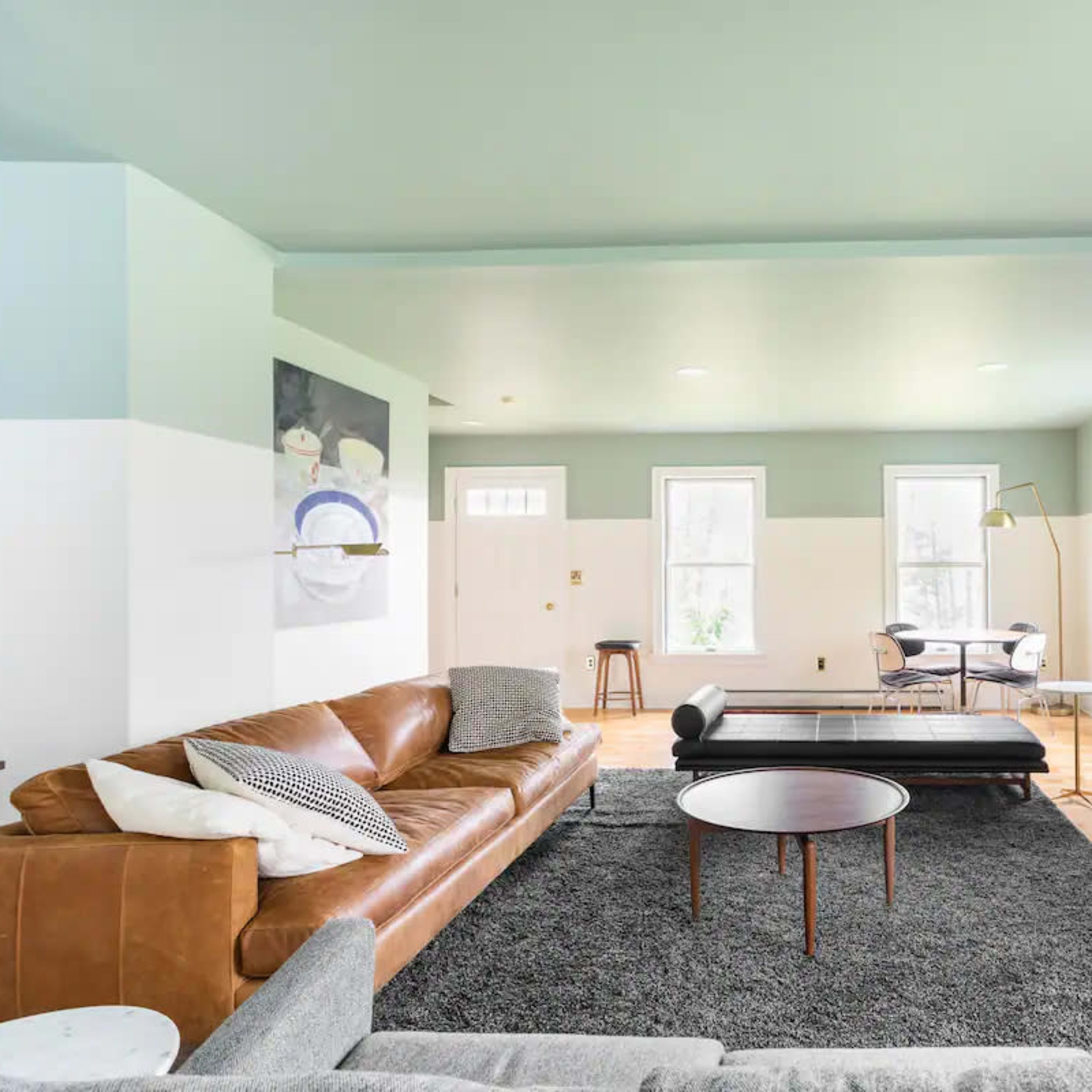 A modern living room features a leather sectional sofa, a round coffee table, a patterned rug, and a fireplace, with large windows allowing natural light.