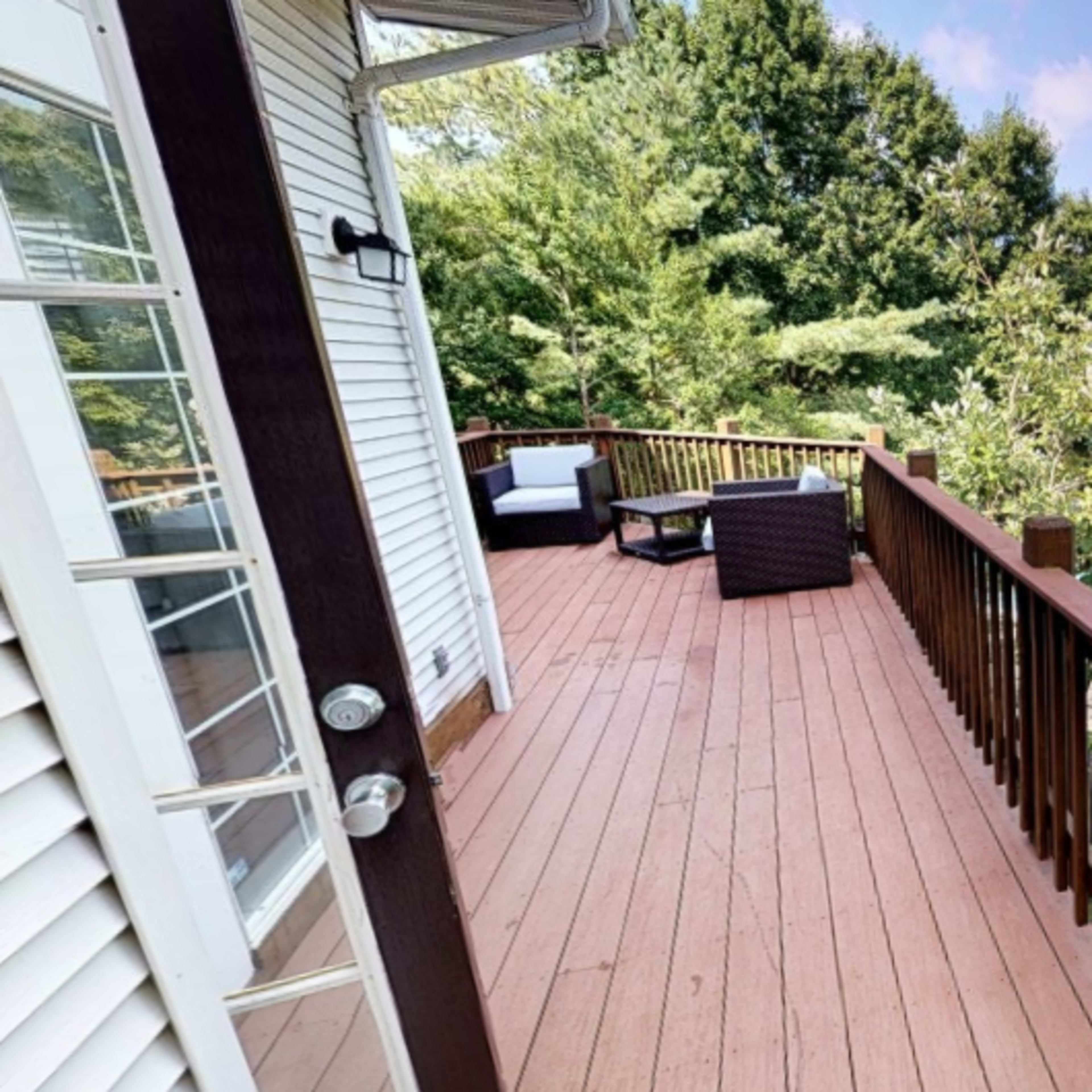 A wooden deck attached to a house, with seating arrangements and a view of greenery in the background.