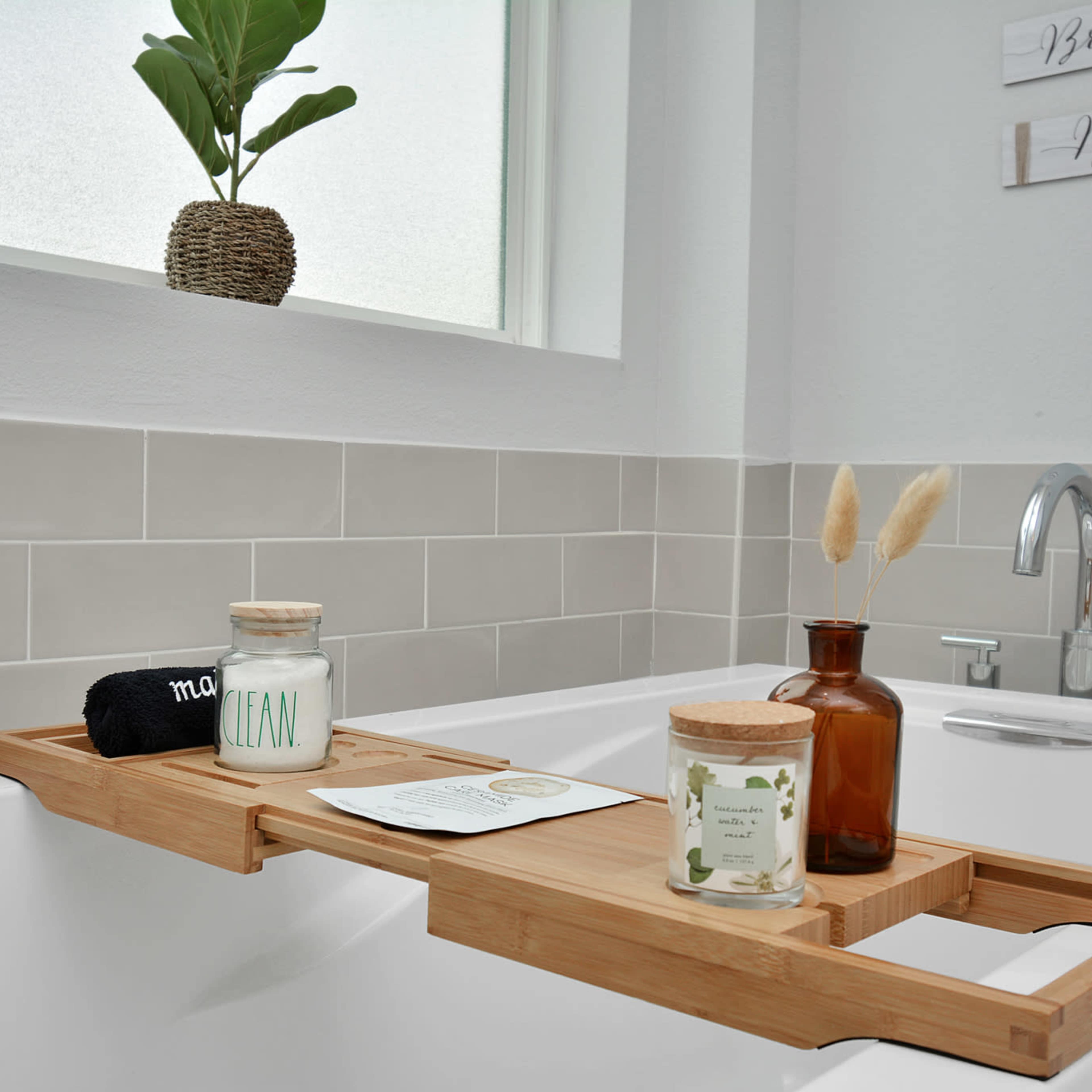 A bamboo bath tray with various candles and a small vase sits across a bathtub next to a window with green plants.