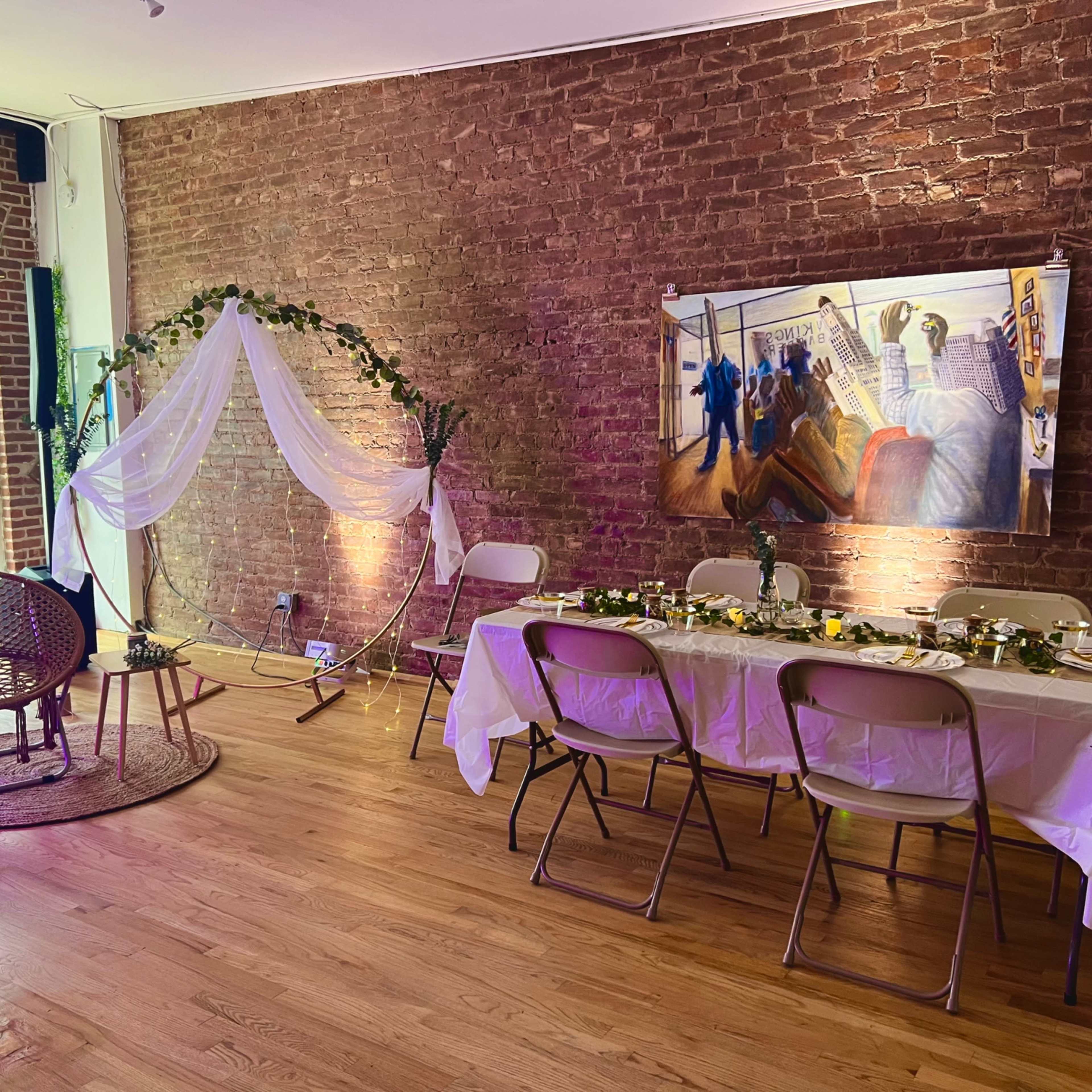 A decorated indoor space featuring a table set for a gathering, a large wicker chair, and a backdrop of an artistic painting on a brick wall.