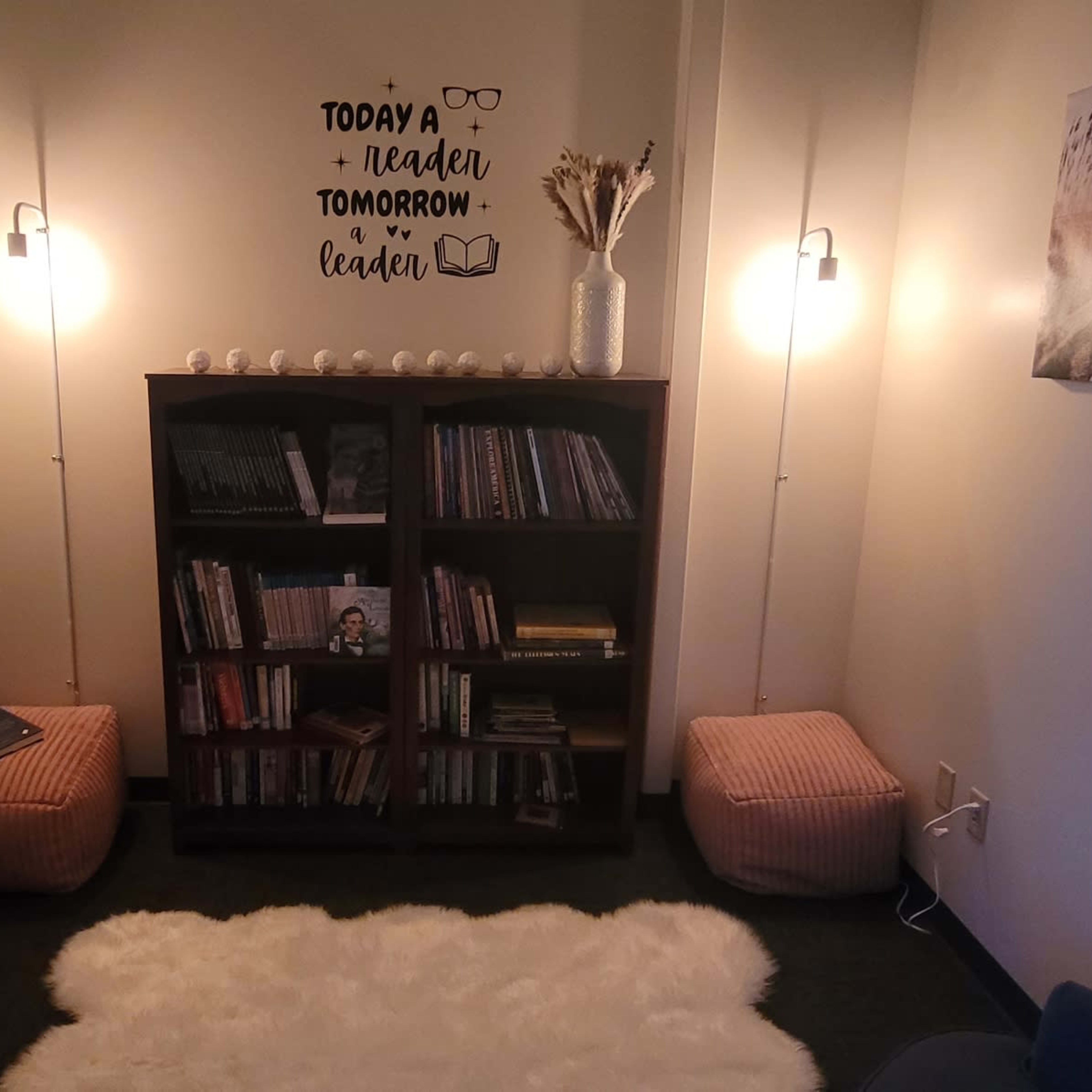 A cozy reading nook with a bookshelf, two poufs, a soft rug, and ambient lighting.