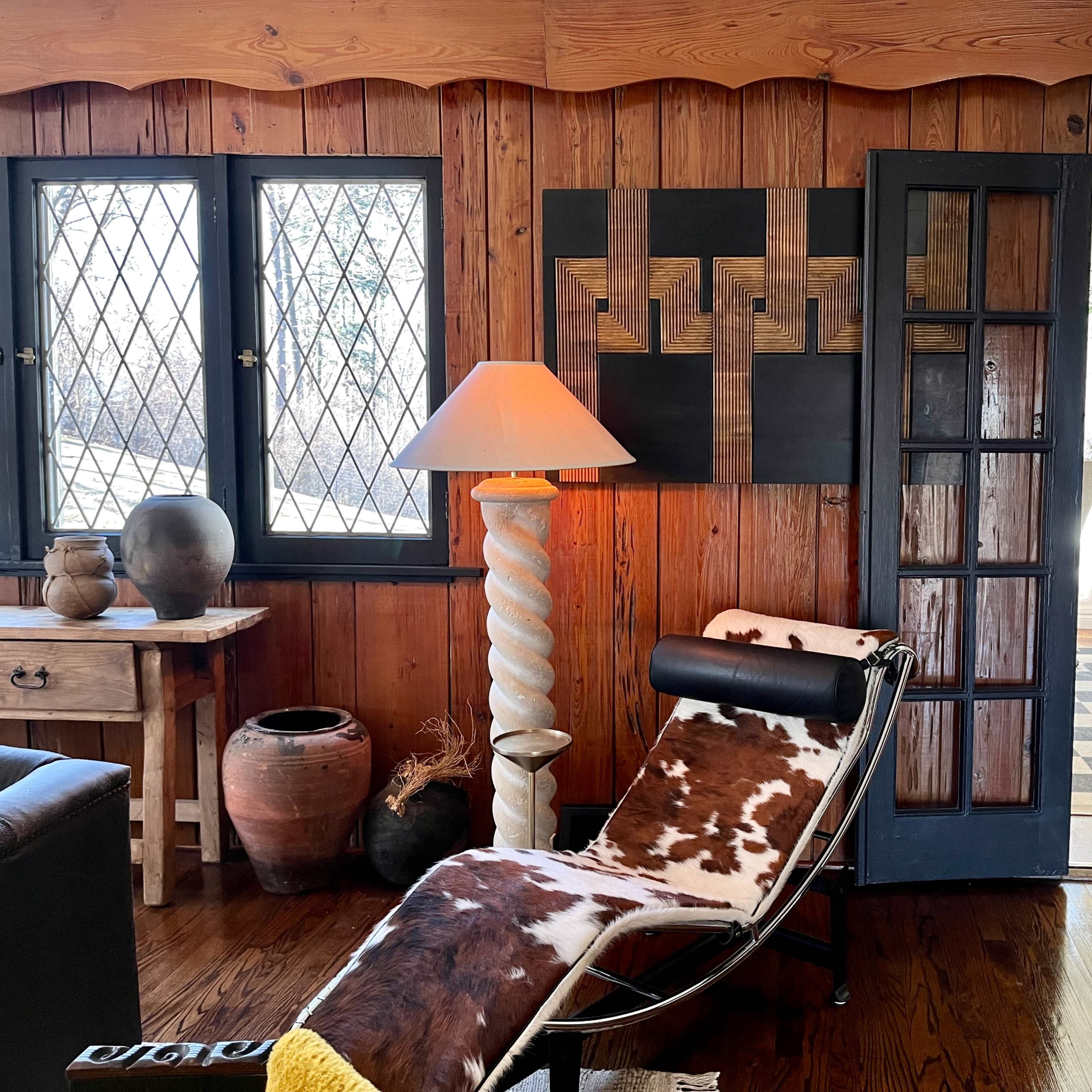A wooden-paneled room with a chaise lounge, a lamp, and a doorway leading to another area featuring plants and patterned flooring.