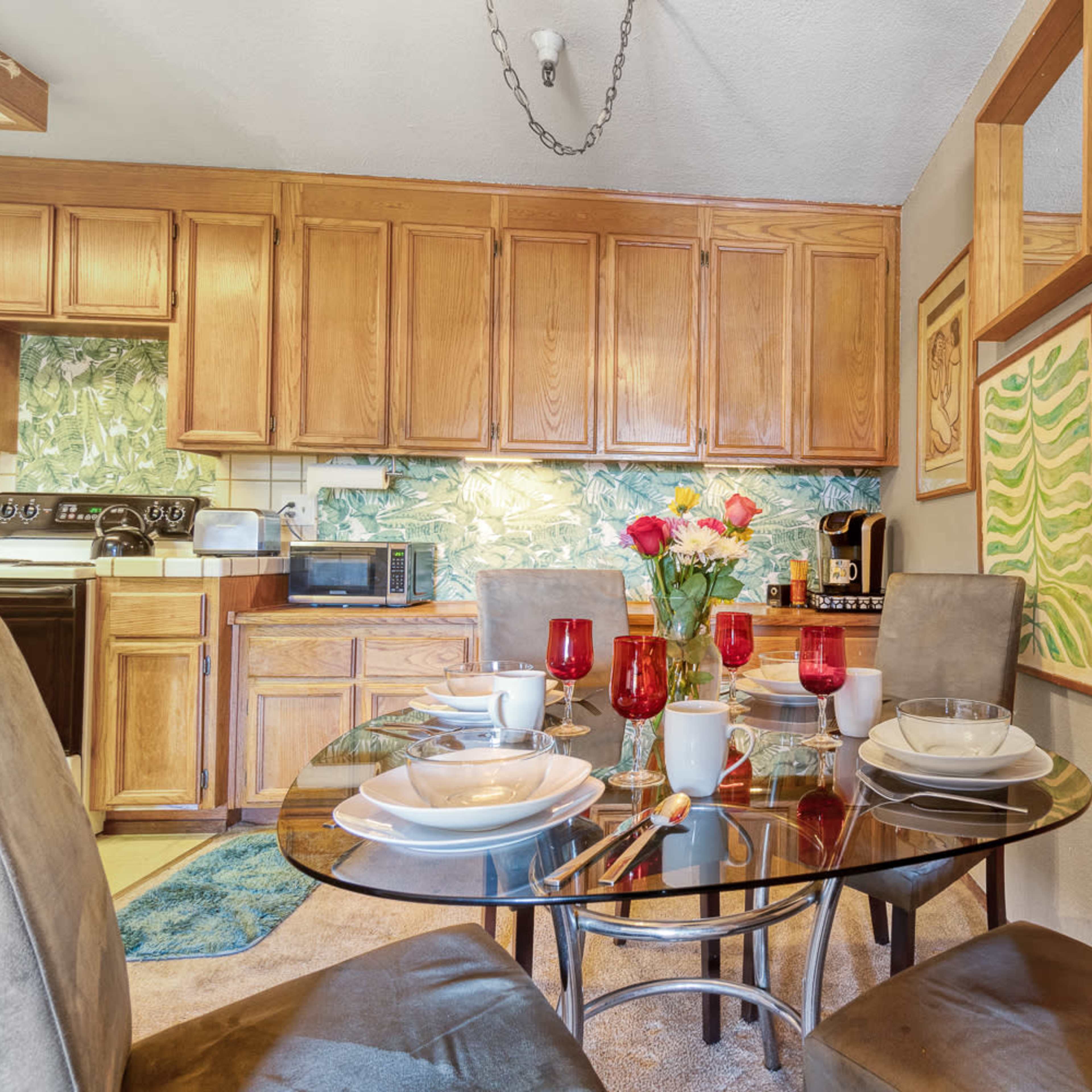 A dining area features a glass table set with dishes and flowers, adjacent to a kitchen with wooden cabinets and decorative wall art.