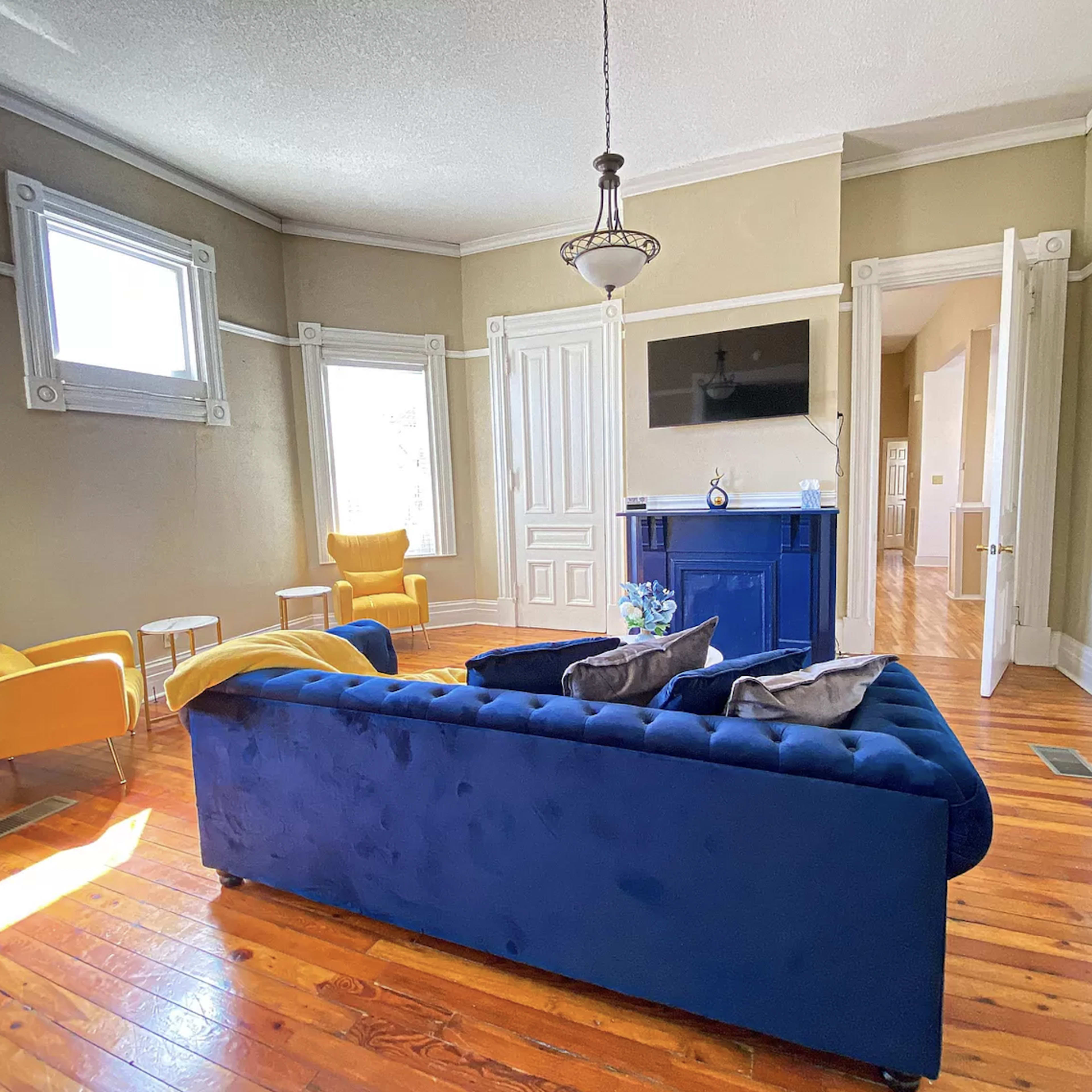 The living room features a blue velvet sofa, yellow chairs, and hardwood flooring, with windows allowing natural light to enter.