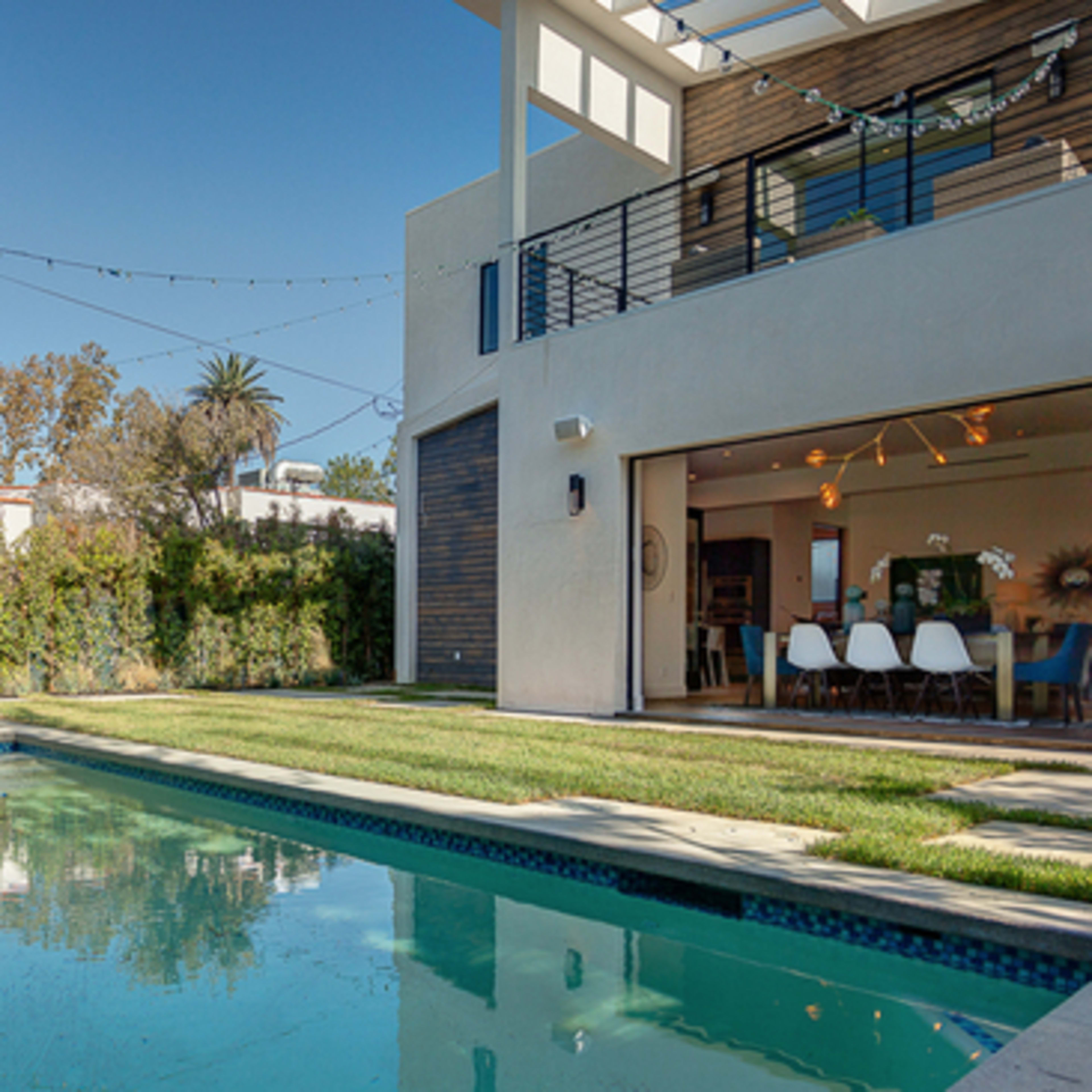 A modern home with a pool and outdoor seating area features large windows that open to a green lawn and surrounding trees.