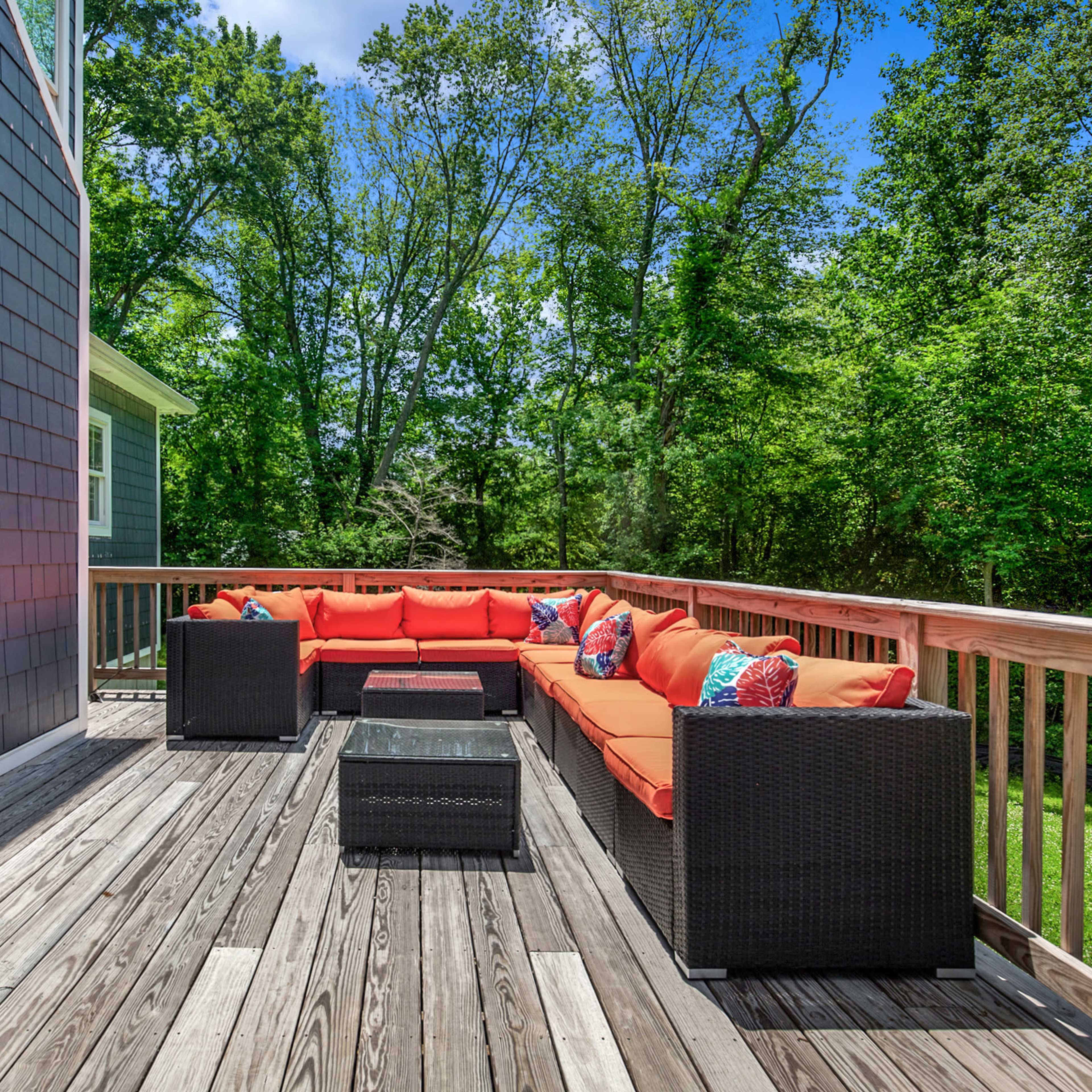 A spacious wooden deck features an orange sectional couch and a black coffee table, surrounded by lush greenery.