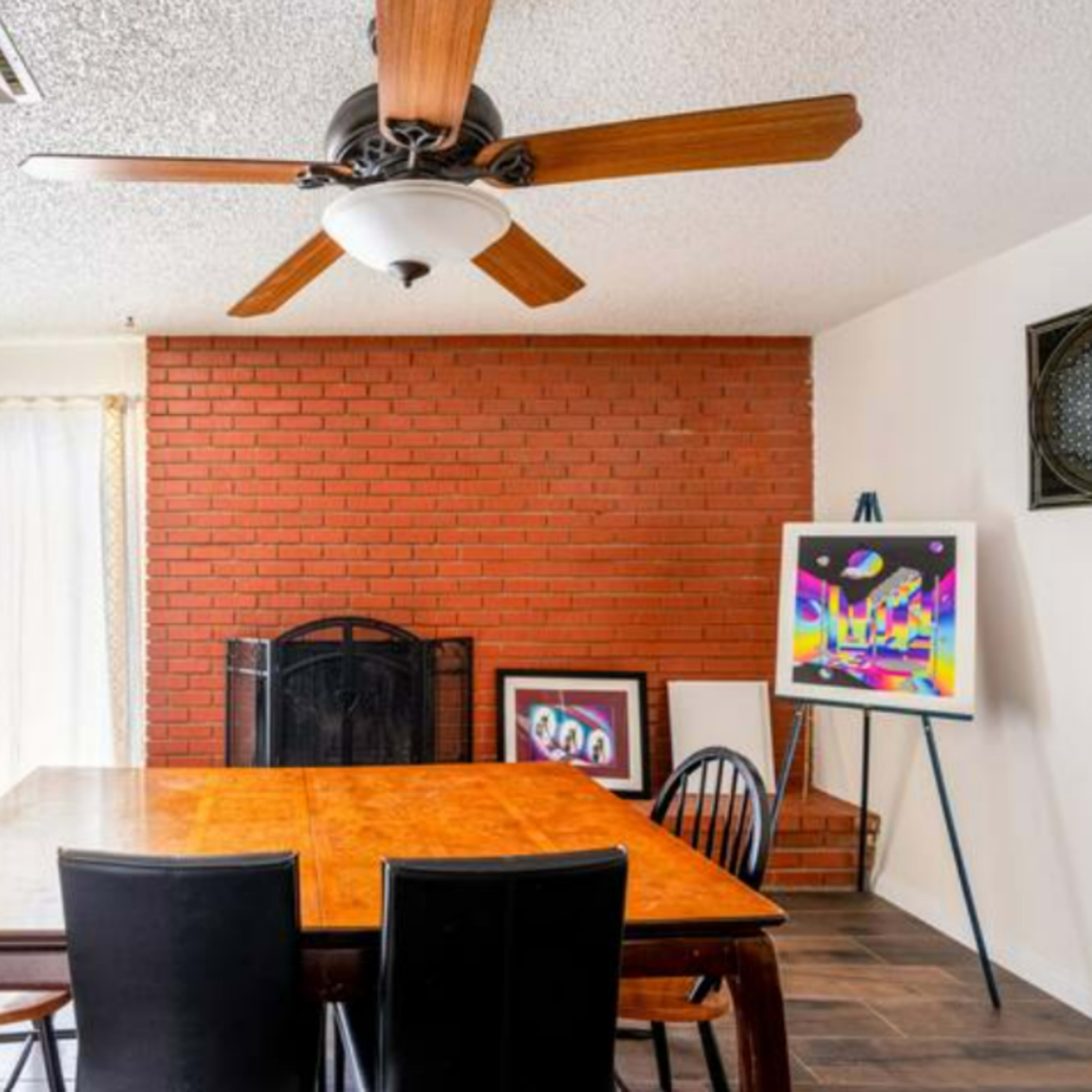 A dining area with a wooden table surrounded by black chairs, a brick wall with a fireplace, and large windows with sheer curtains.