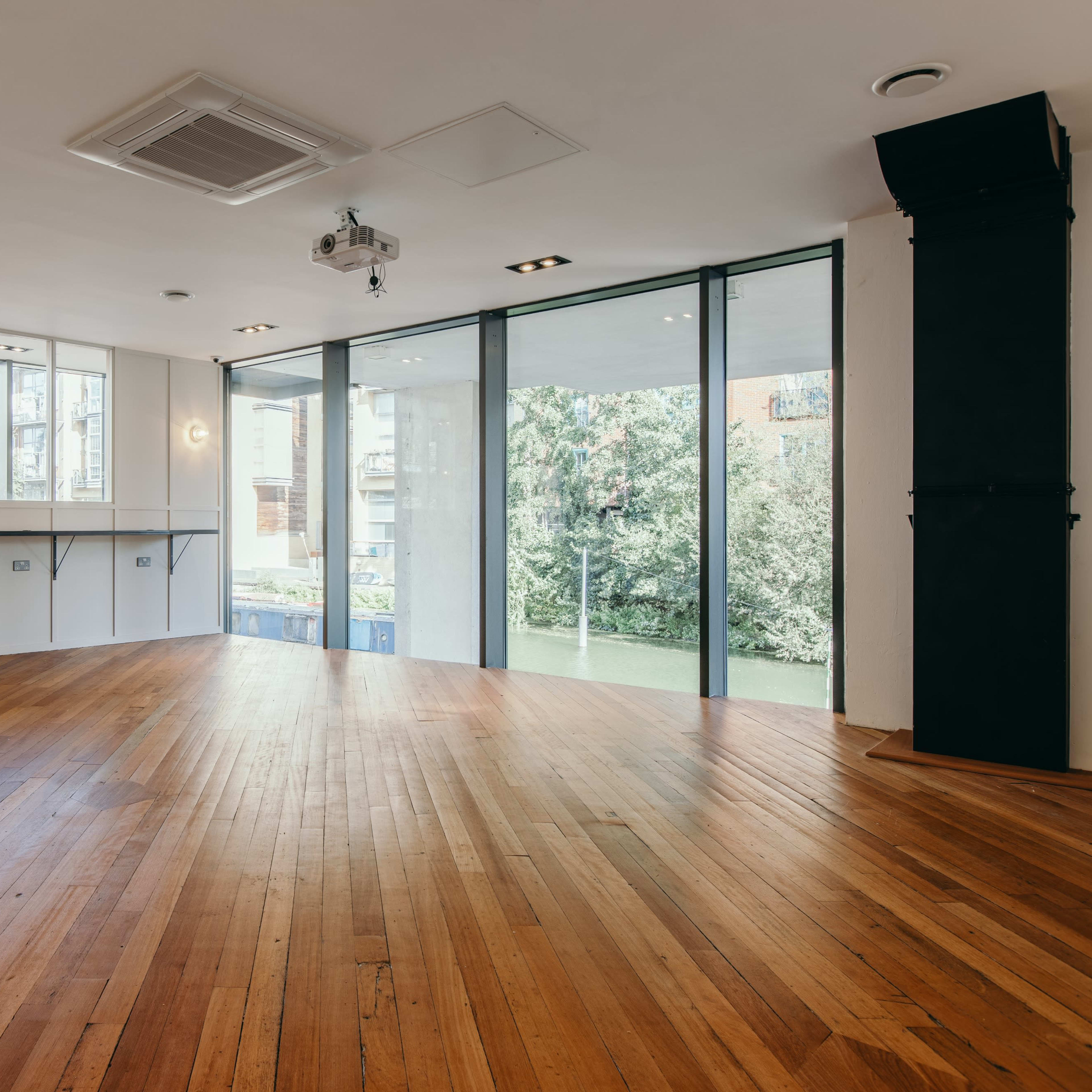 An empty room with wooden flooring and large glass windows overlooking a garden.