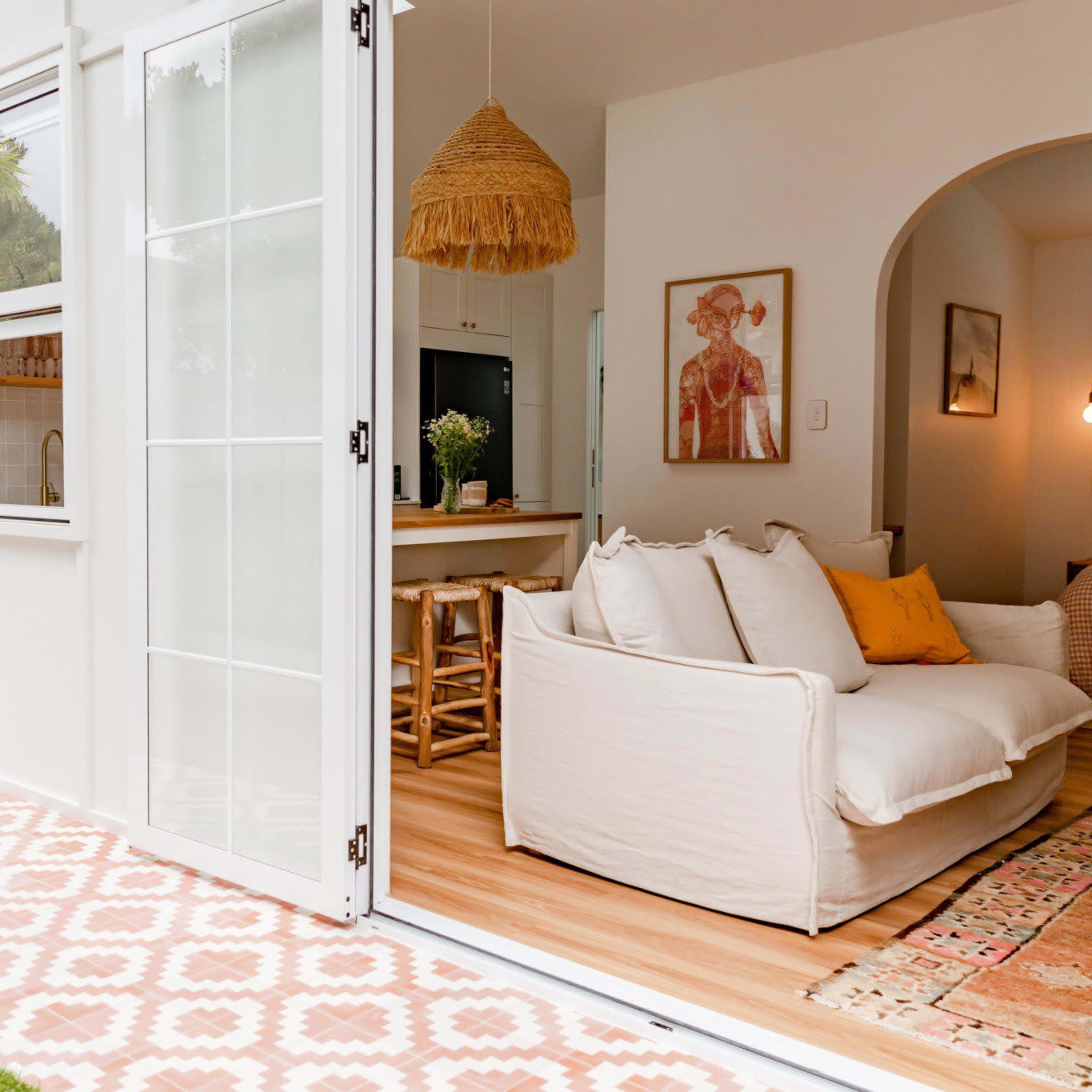 A bright living space features an open door leading to a patio with laid tile and a green lawn, showcasing a cozy couch and a kitchen area in the background.