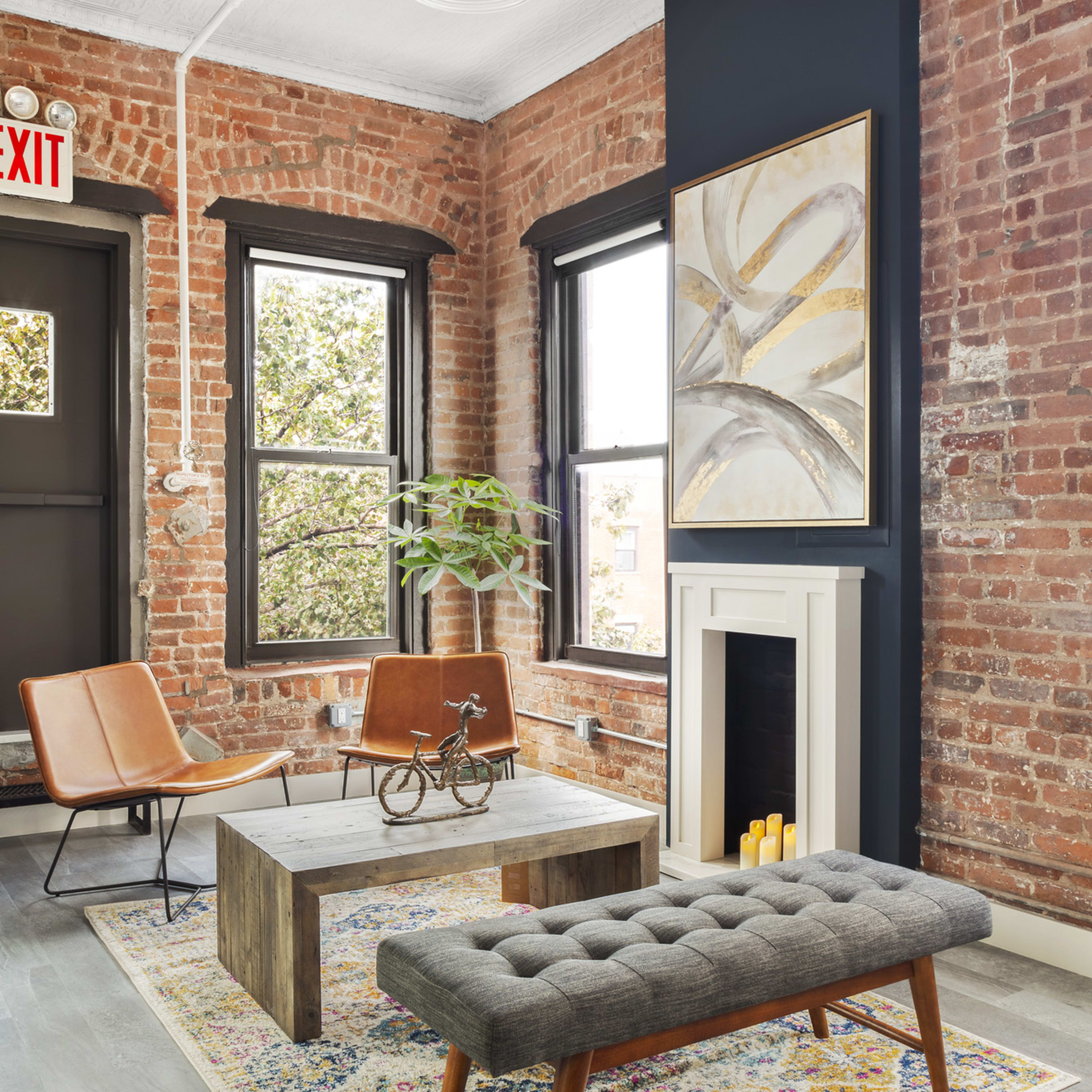 A cozy living room with exposed brick walls, featuring two modern chairs, a wooden coffee table, a tufted bench, and a decorative rug.