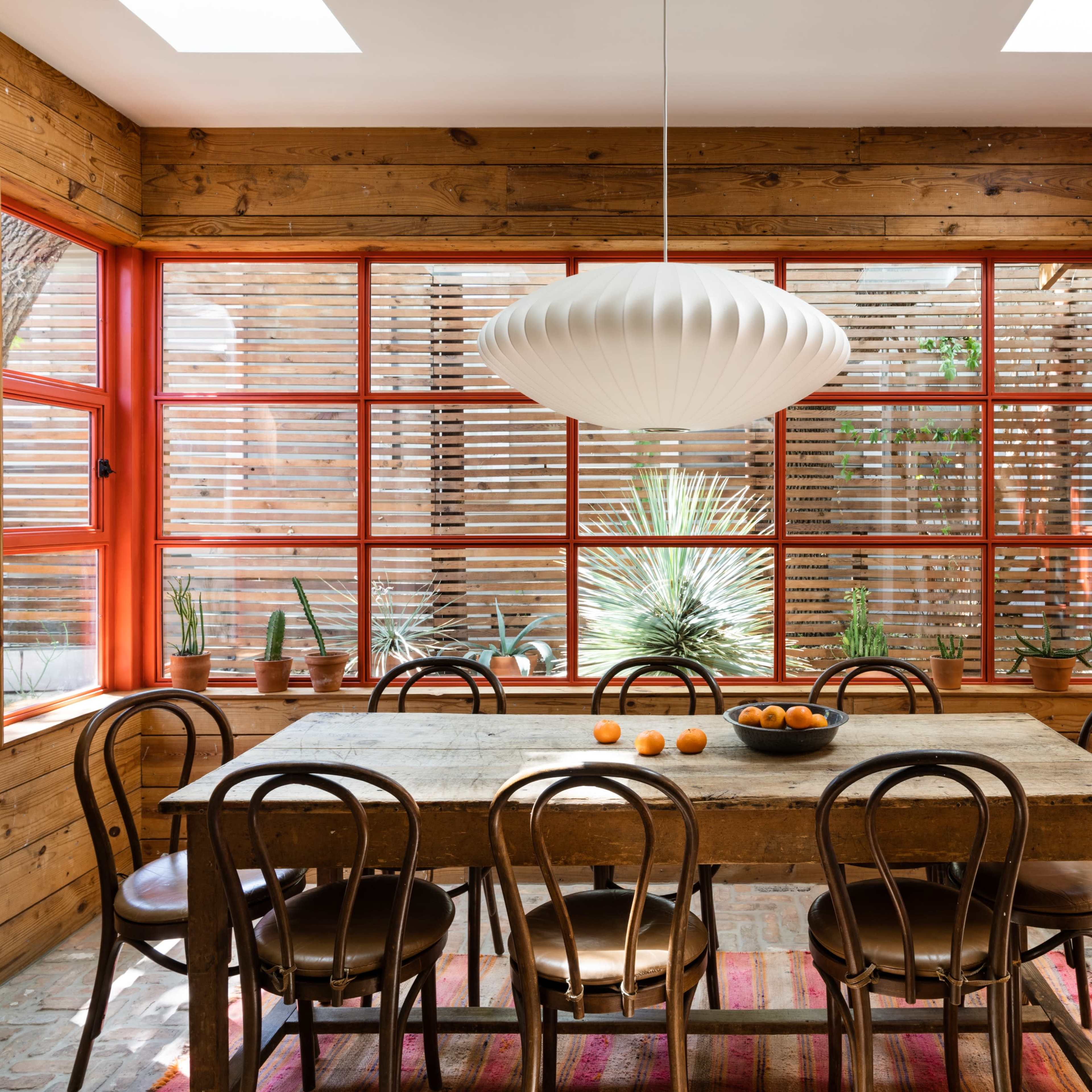 A wooden dining table surrounded by chairs is situated in a bright room with large windows and orange trim, featuring a bowl of oranges in the center.