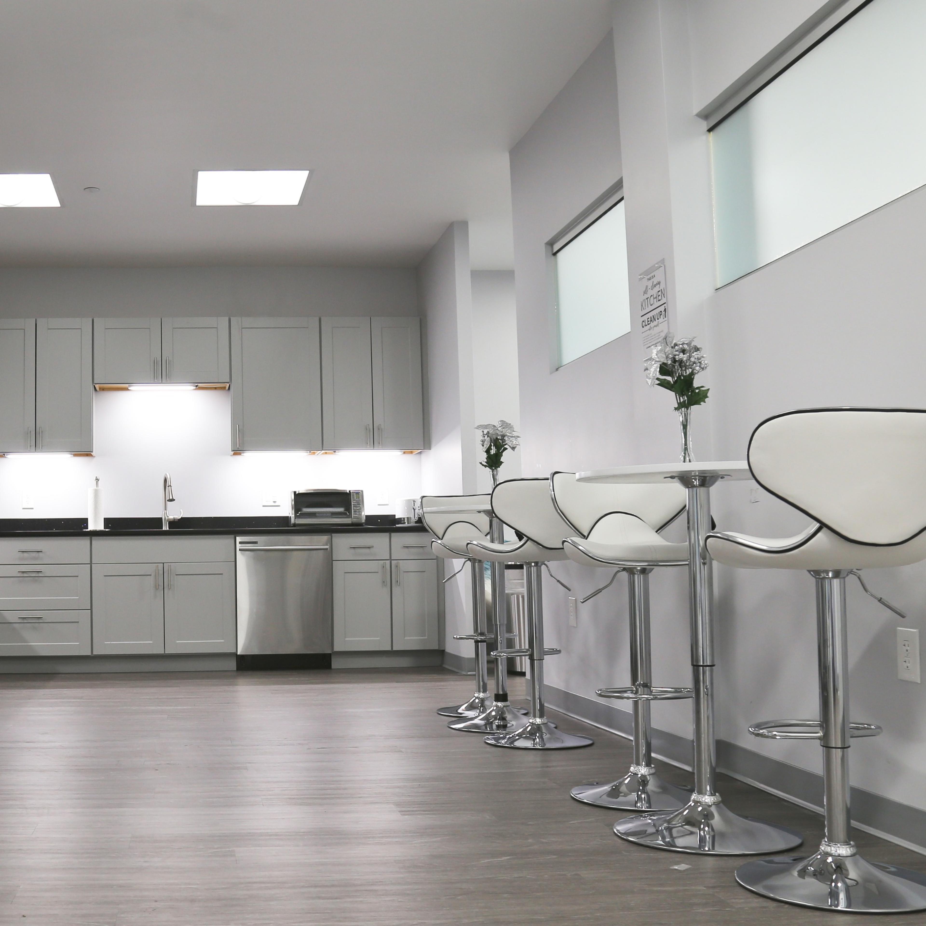 A modern kitchen with white cabinets, stainless steel appliances, and a row of white bar stools along a counter.