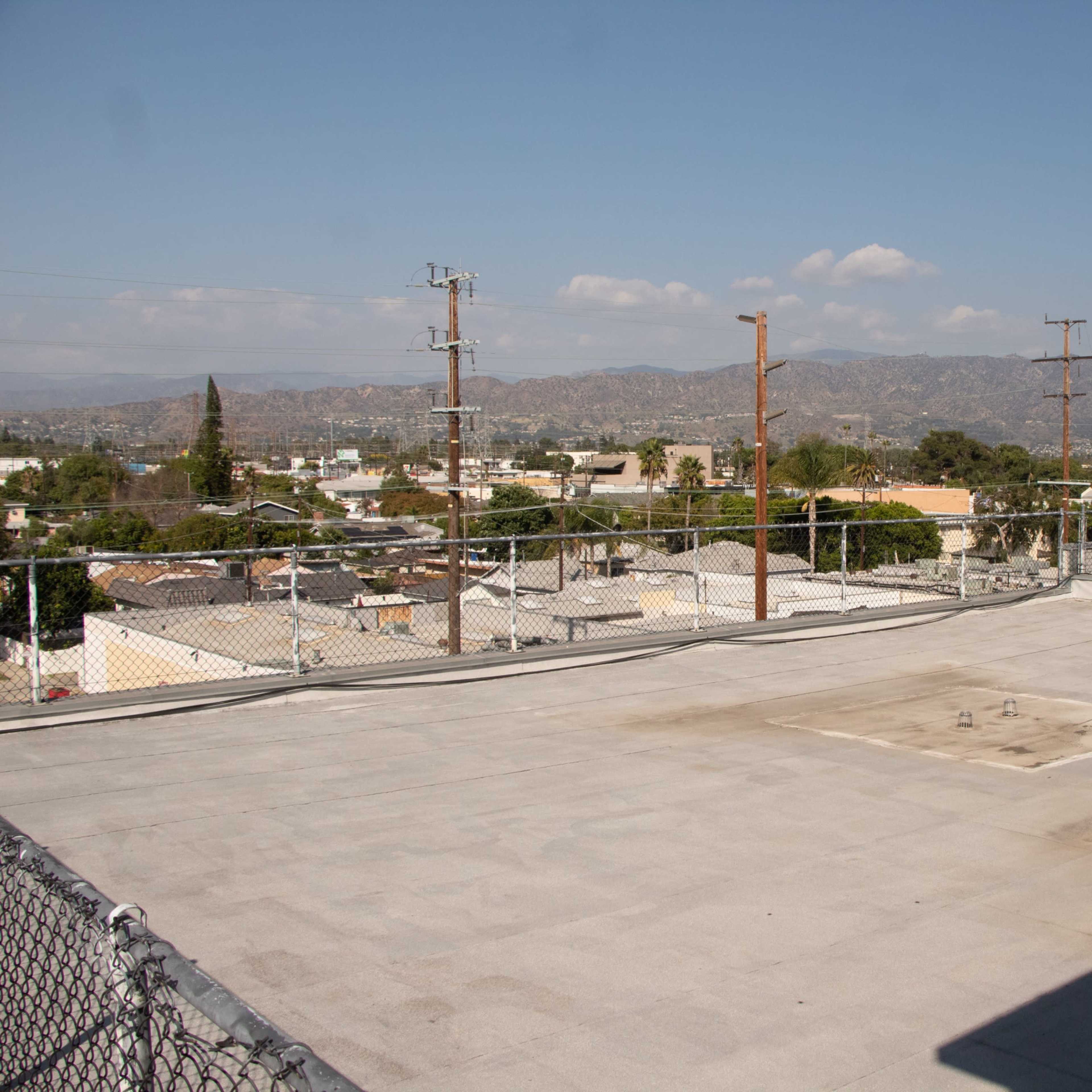 A flat rooftop with a fenced perimeter overlooking a cityscape and distant mountains under a clear sky.