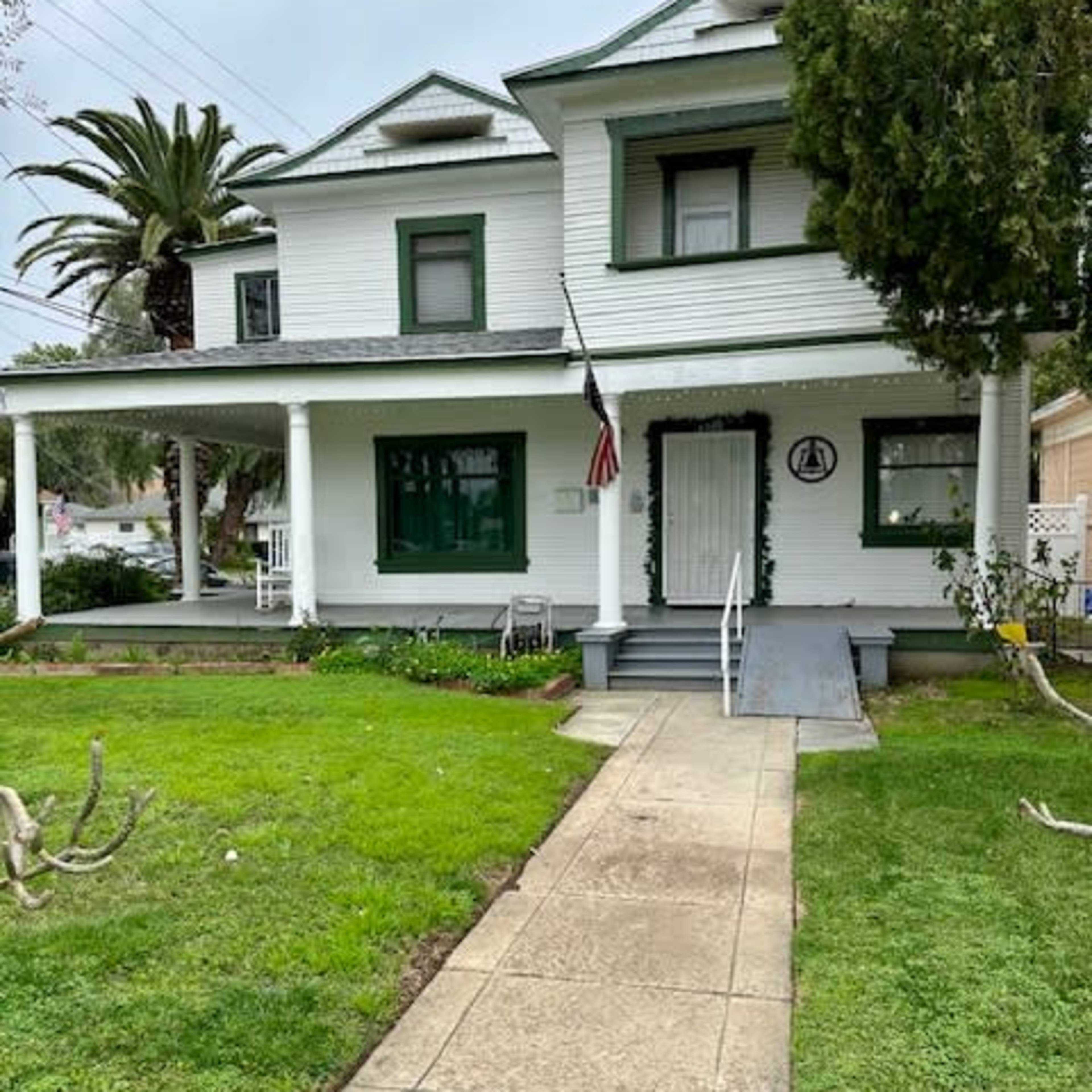 A white two-story house with green trim features a front porch, a pathway leading to the door, and palm trees in the yard.