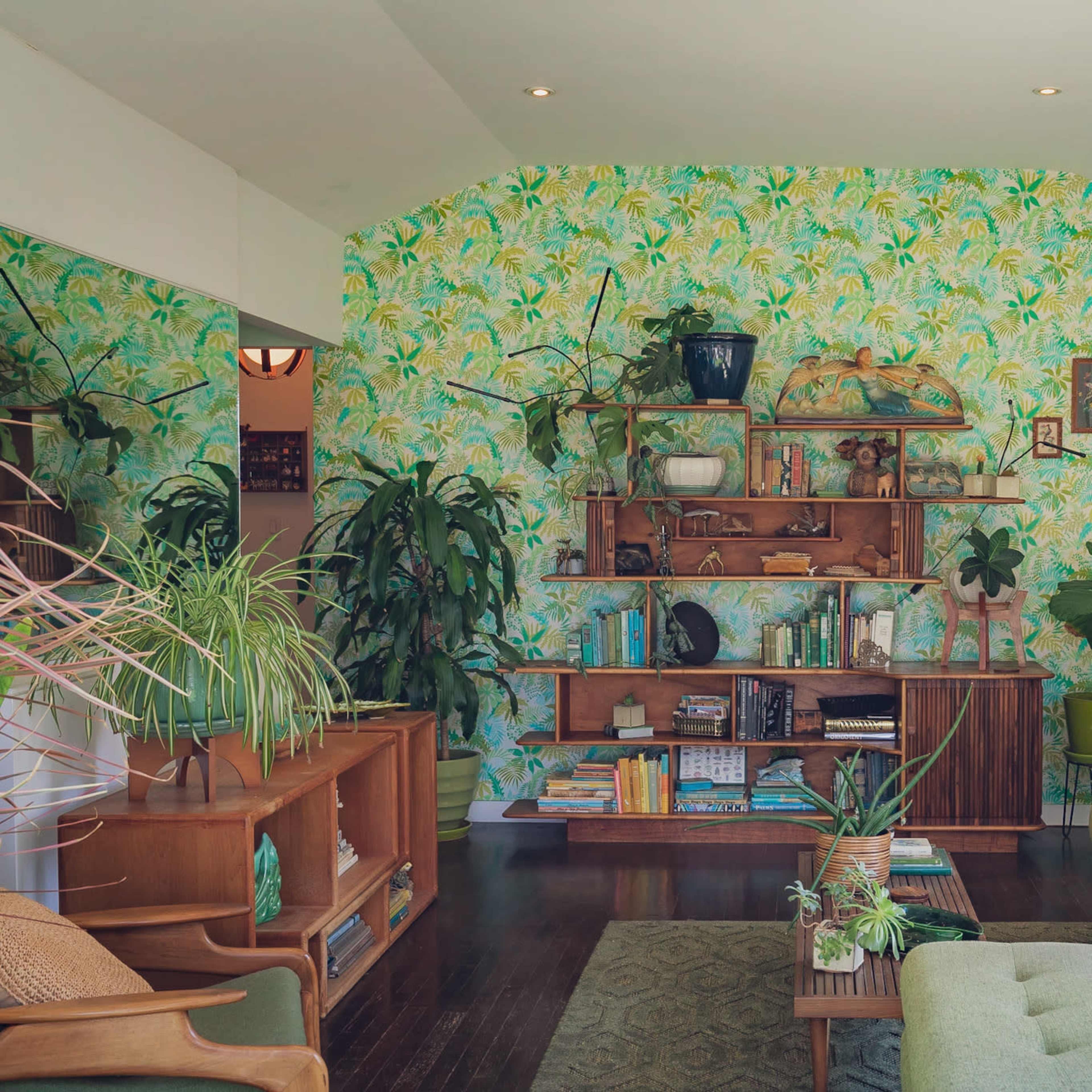 The living room features a green sectional sofa, a wooden bookshelf filled with books and plants, and a wall covered in tropical-patterned wallpaper.