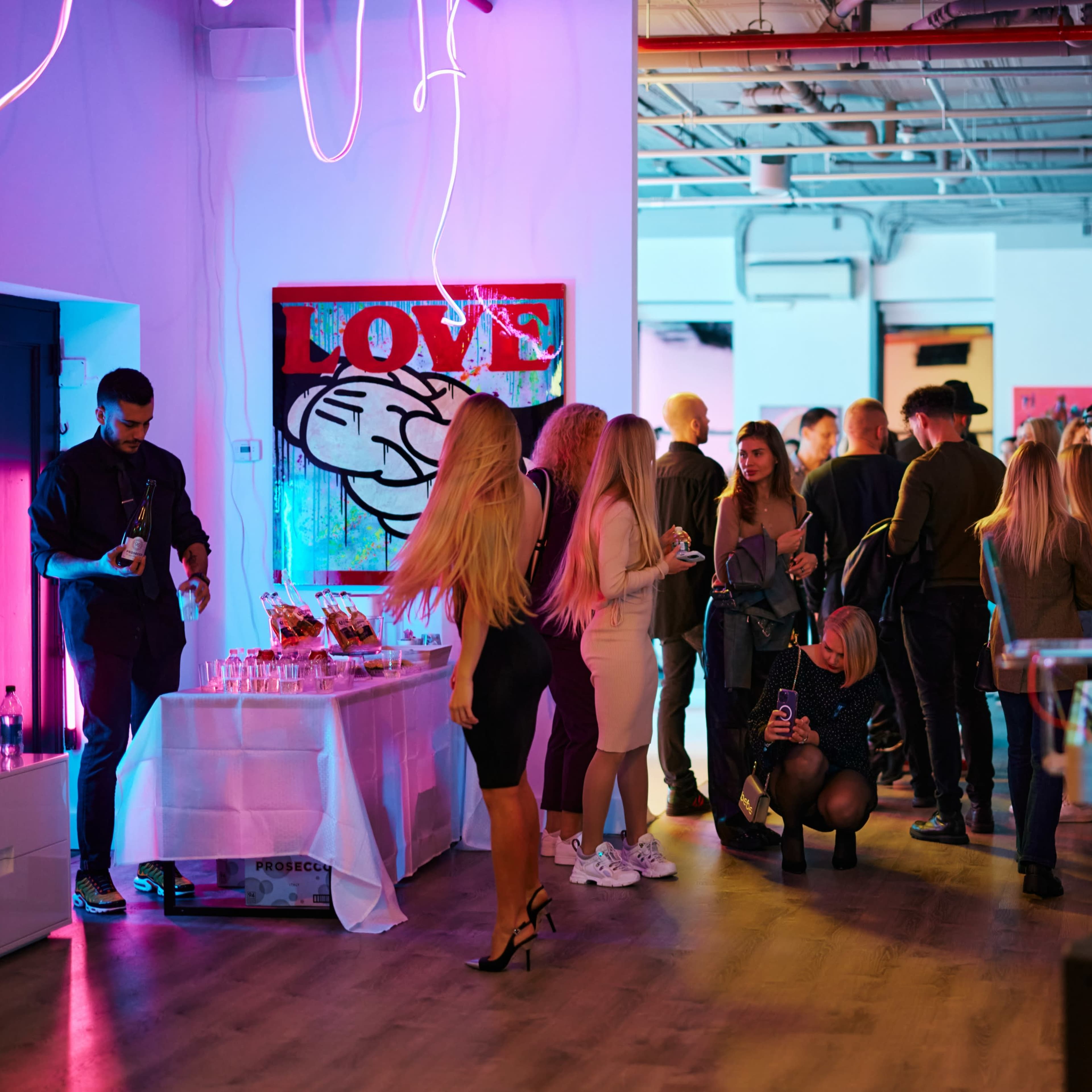 A lively art gallery event features guests mingling, with a DJ playing music and colorful neon lights illuminating the space.