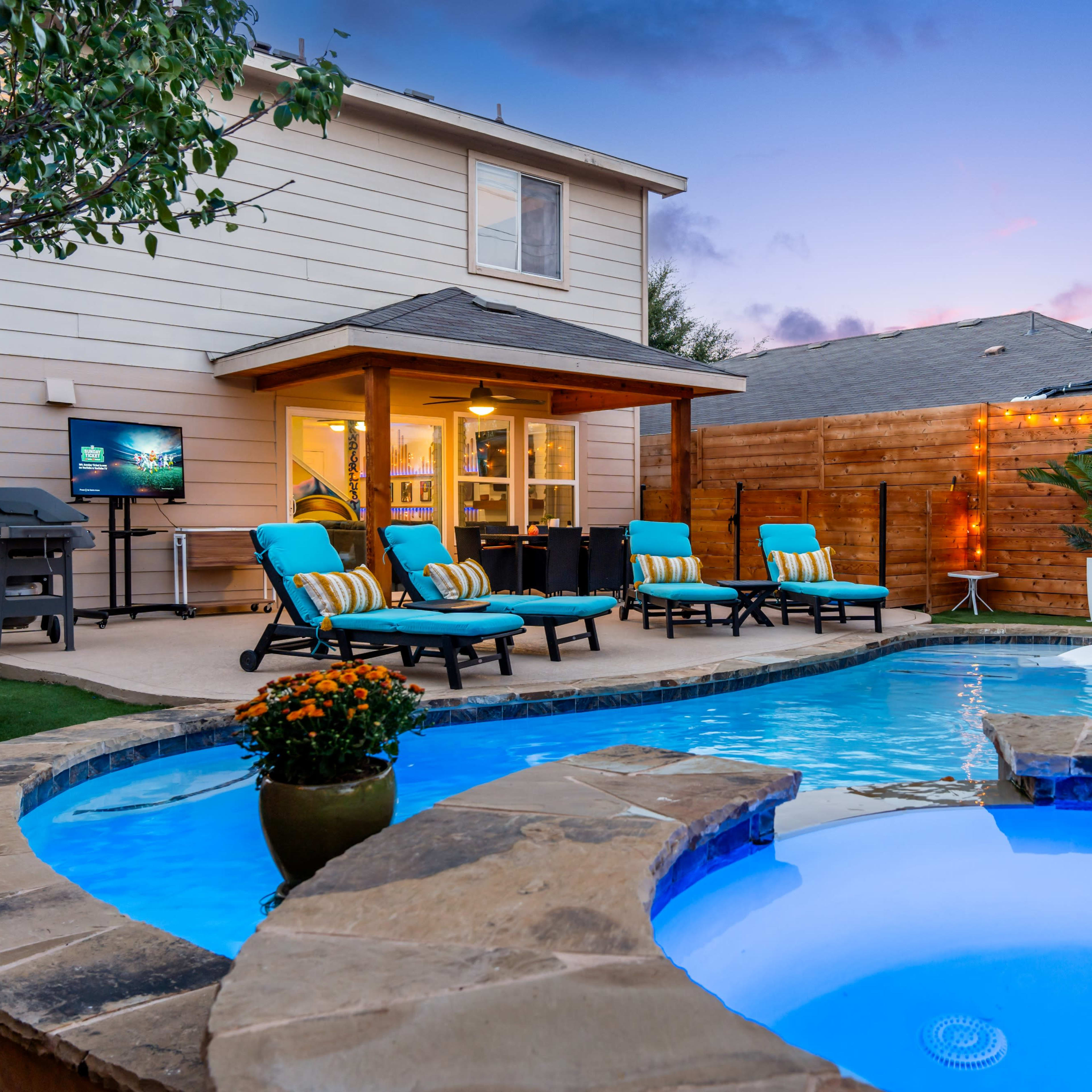 A backyard with a swimming pool, lounge chairs, and a small garden area, framed by a house and wooden fencing under a colorful sky.
