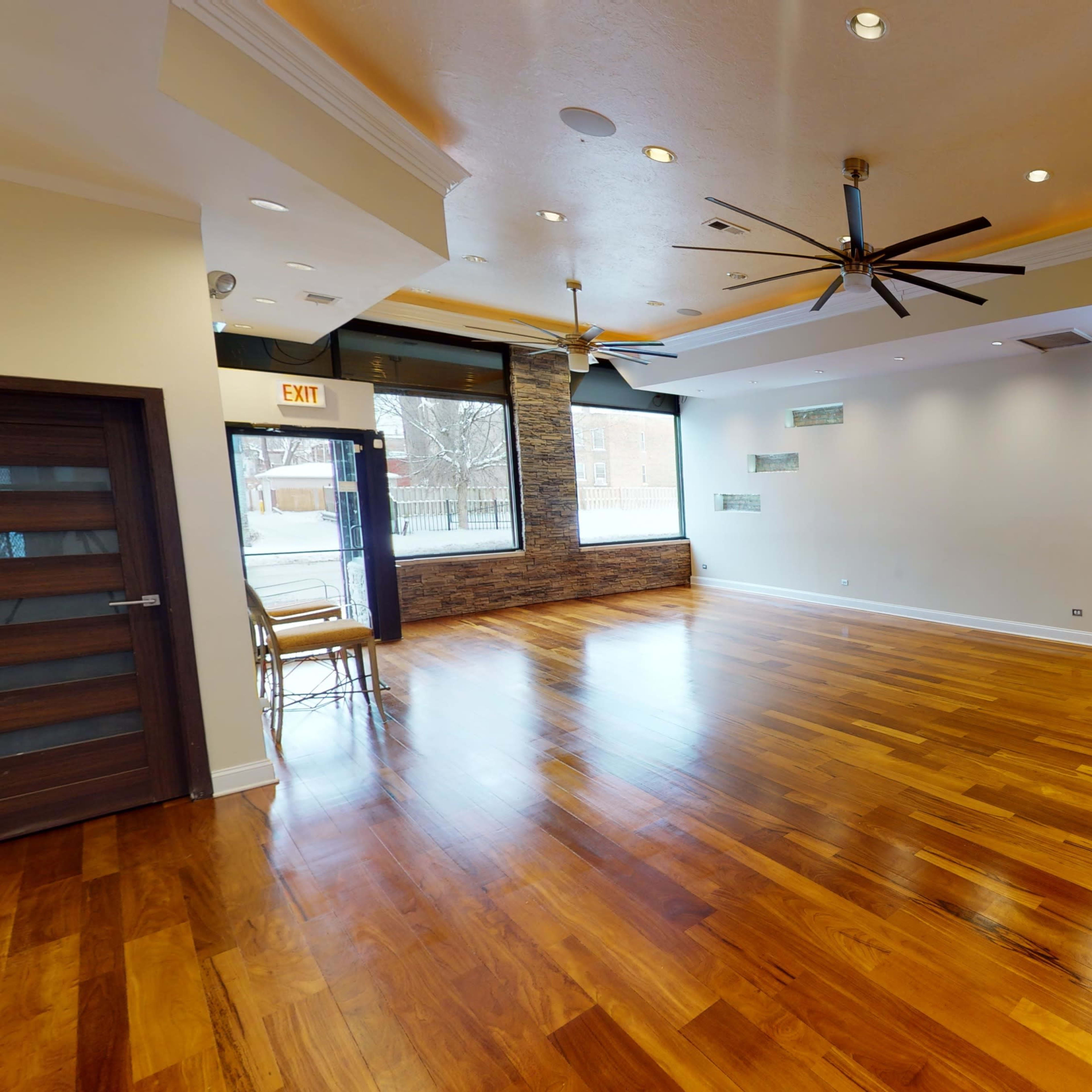 A spacious room with hardwood flooring, large windows, and ceiling fans.
