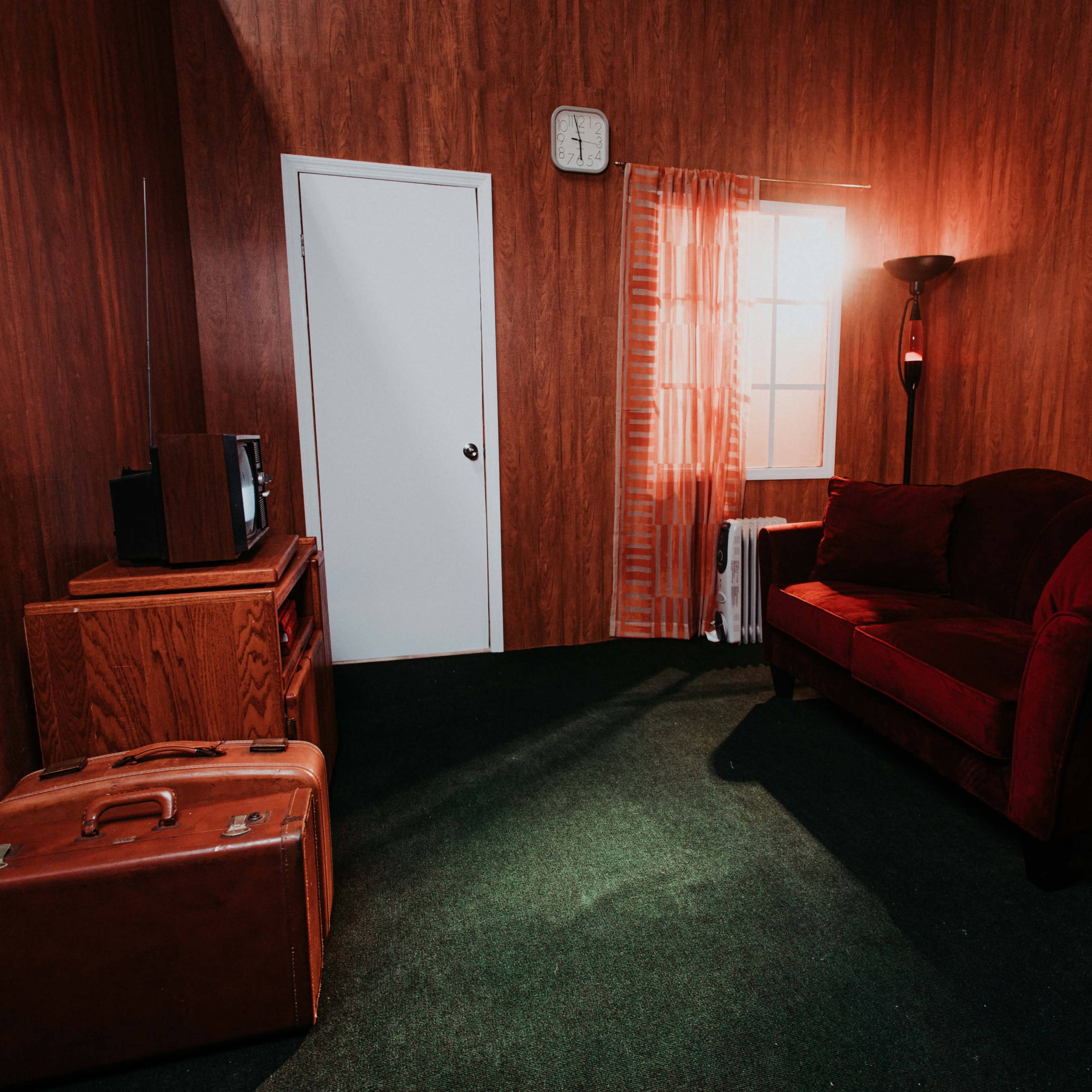 A small room with wooden walls, a red couch, an old television on a wooden cabinet, a suitcase on the green carpet, and a window with sheer curtains letting in light.