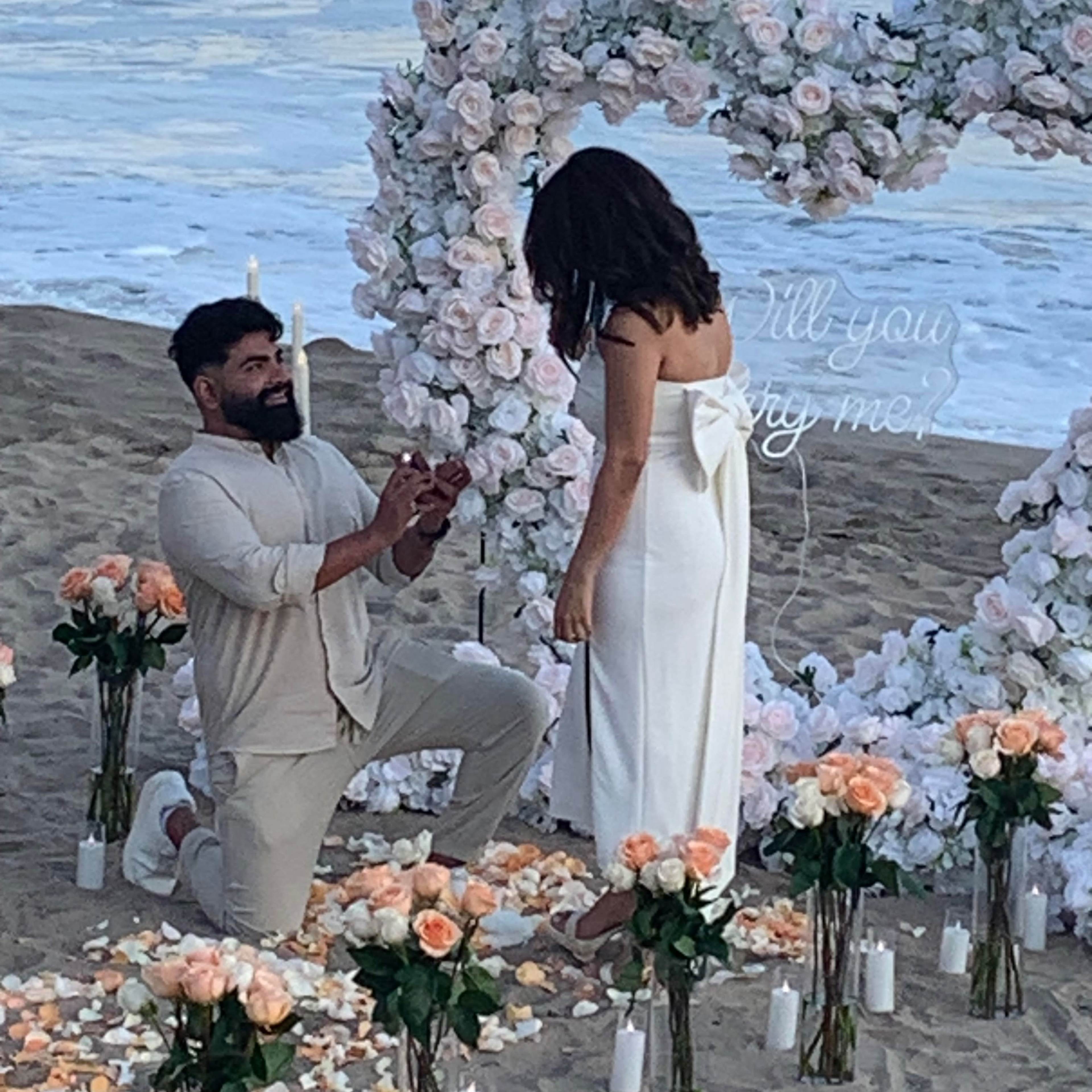 A man is proposing to a woman on the beach, kneeling in front of a floral heart arrangement with candles and rose petals scattered around them.
