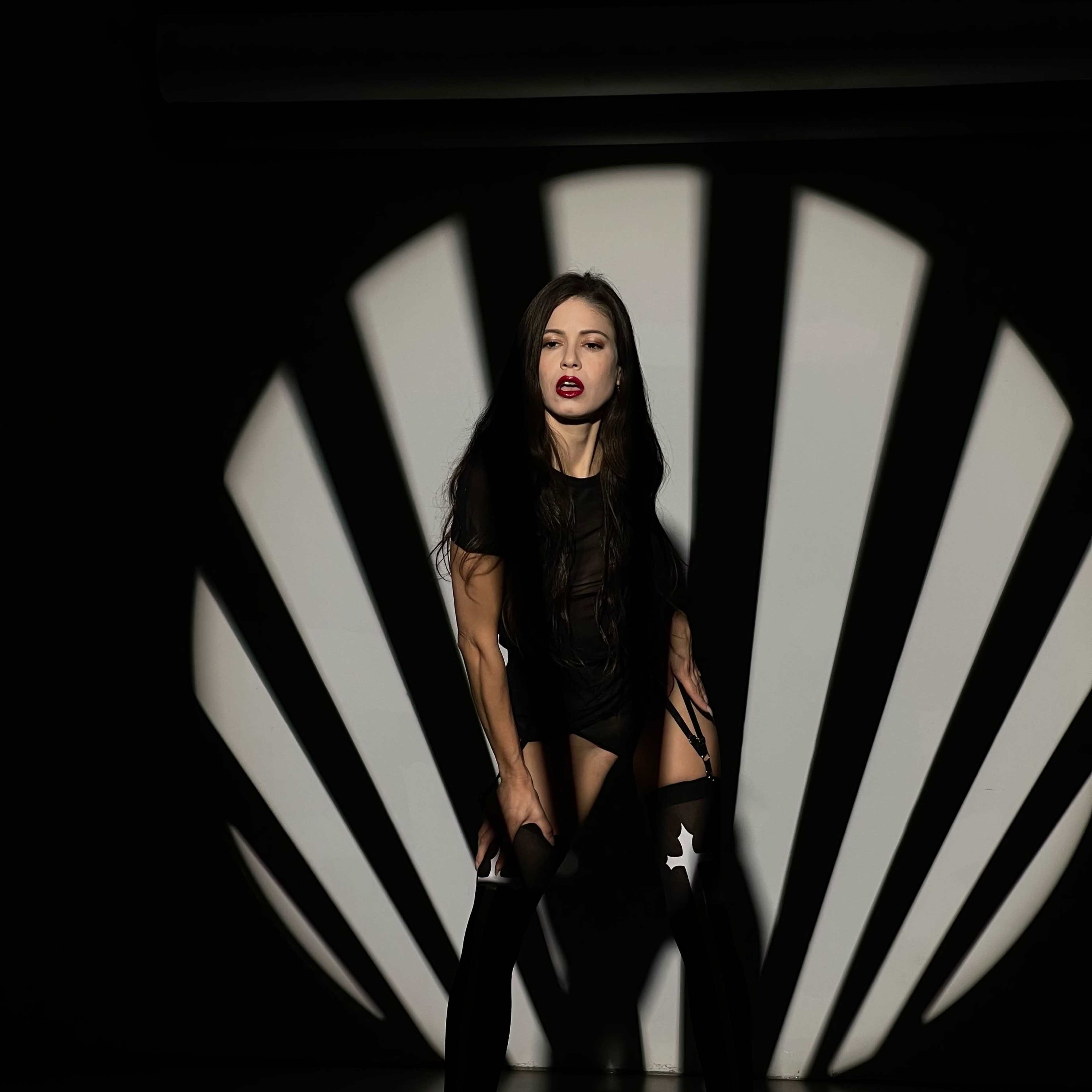 A person in a dark outfit poses against a backdrop of radial shadow patterns on the wall.