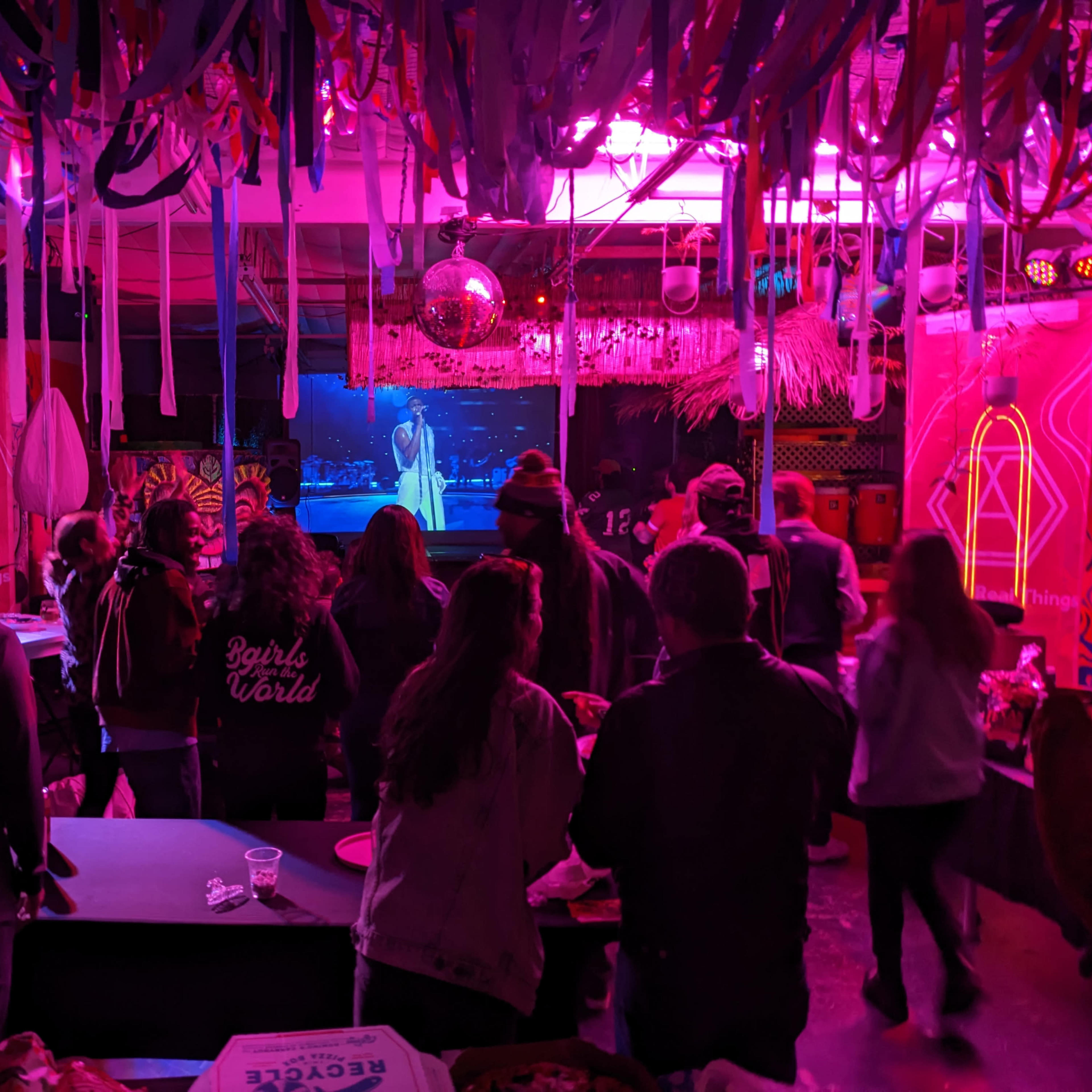 A group of people watches a performer on stage in a colorful venue adorned with streamers and vibrant decorations.