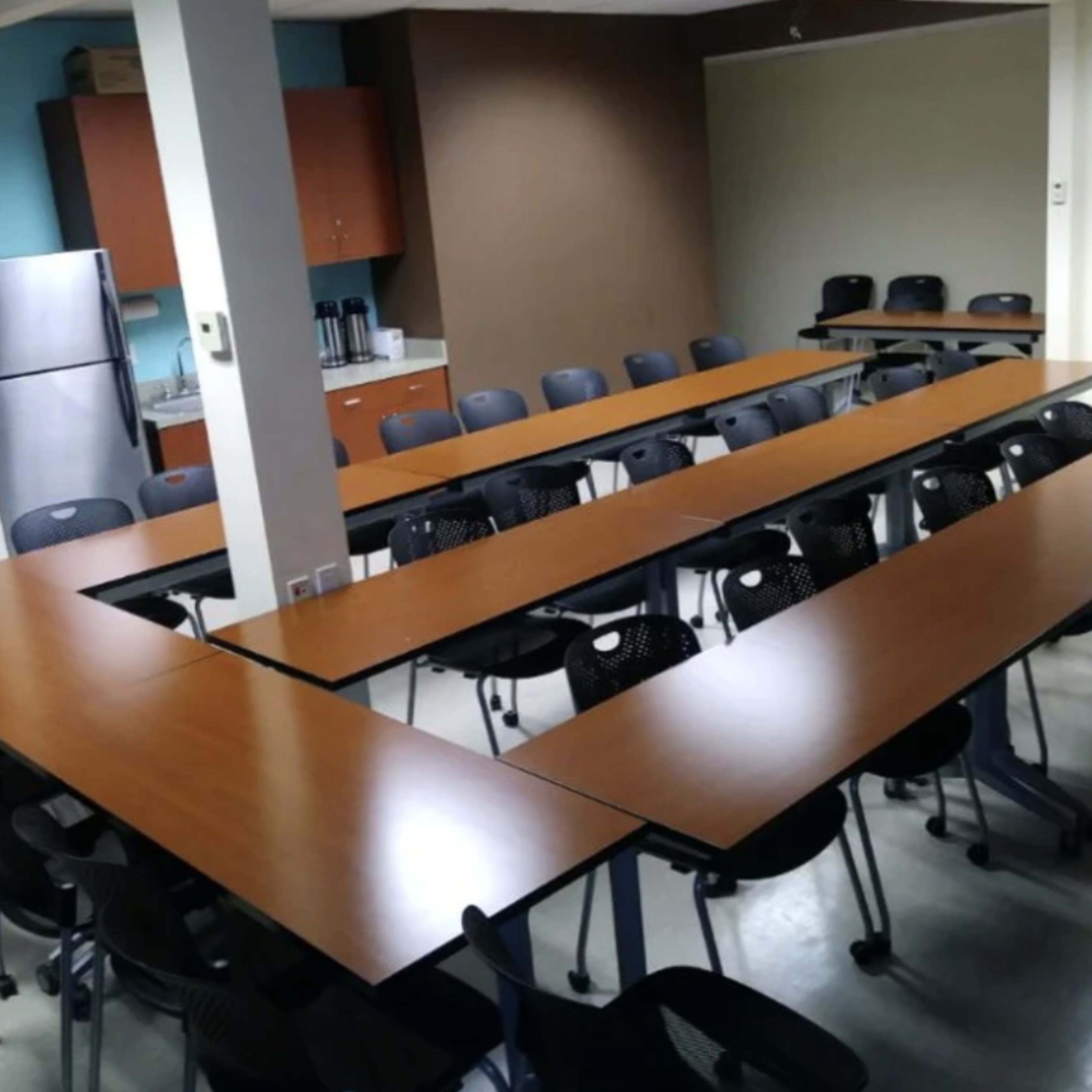 A conference room is set up with multiple long tables arranged in a U-shape, surrounded by chairs, and equipped with a kitchenette in the background.