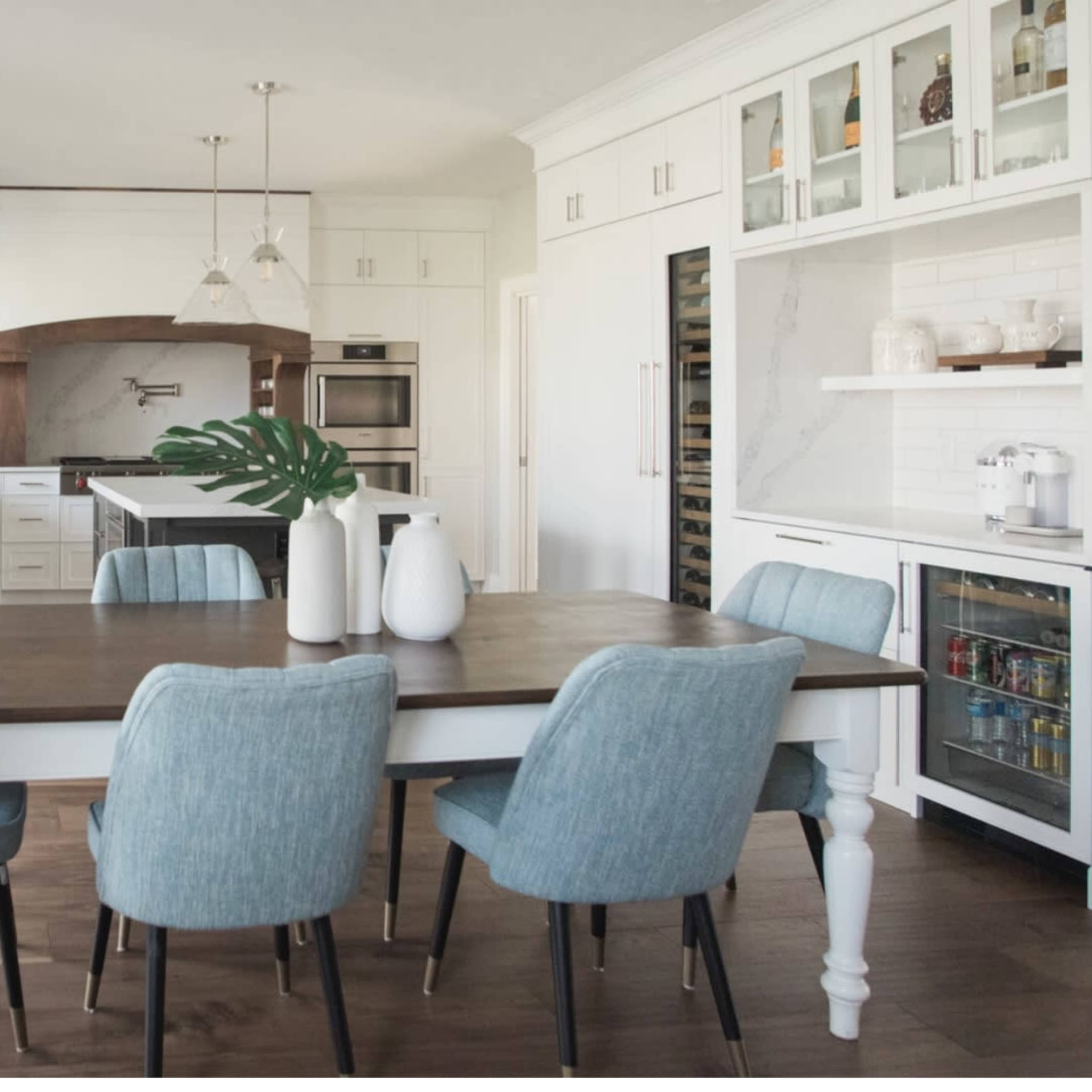 A modern kitchen with a wooden dining table surrounded by light blue upholstered chairs, and cabinetry featuring white and wood accents.