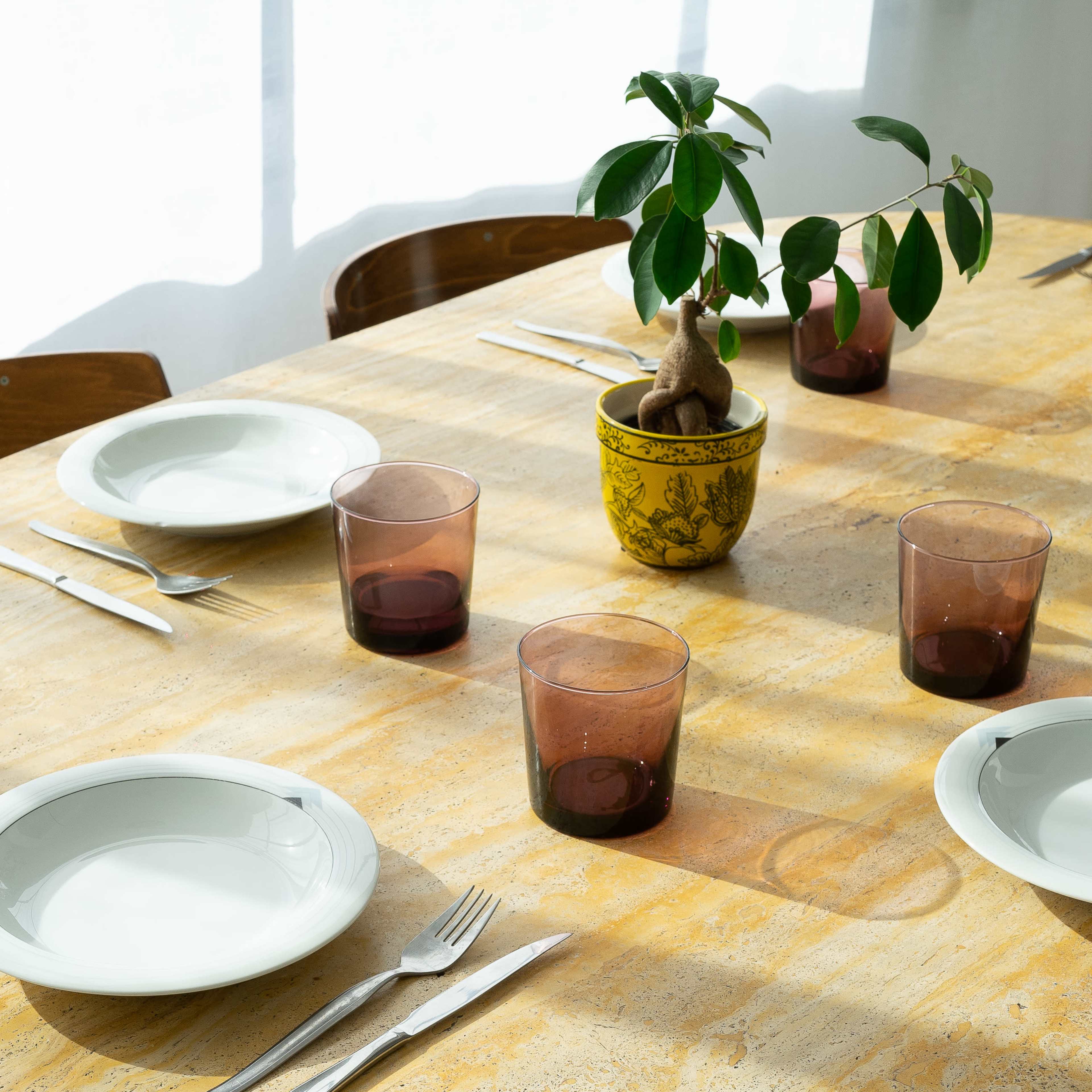 A round dining table is set with white plates and brown glasses, surrounded by wooden chairs and a potted plant in the center.