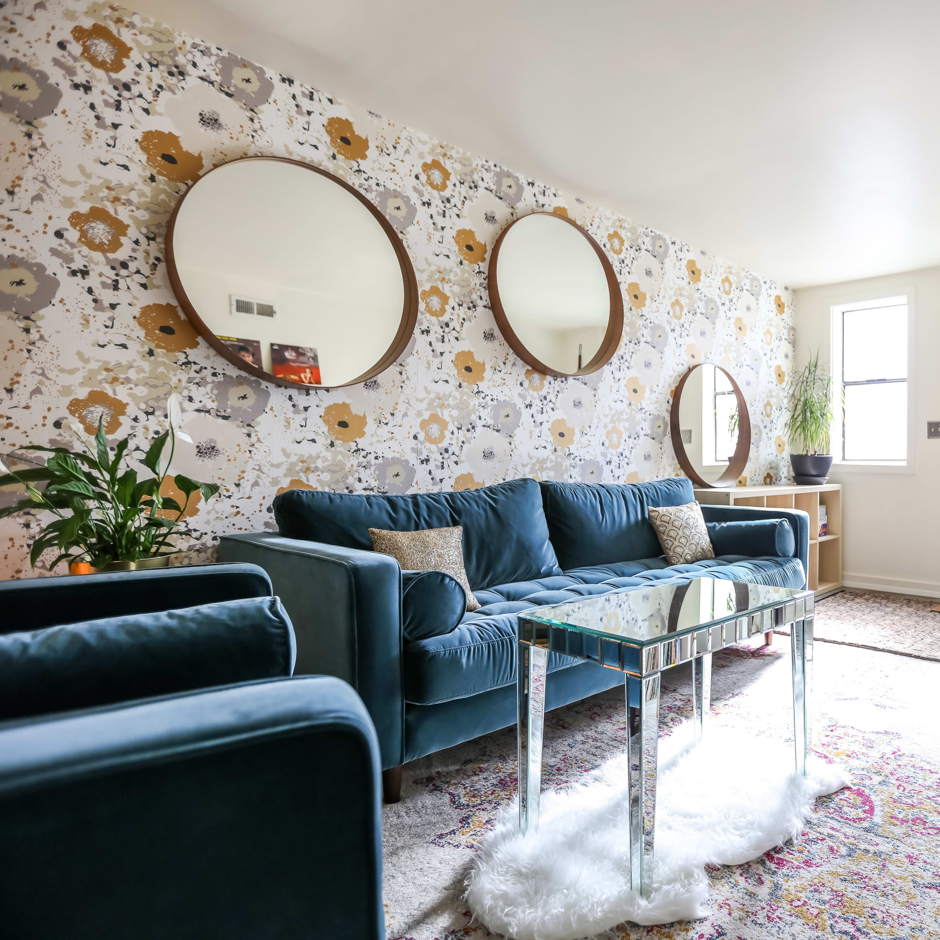 A well-decorated living room featuring a teal sofa, circular mirrors on the floral wallpaper, and a glass coffee table near a door with a small window.
