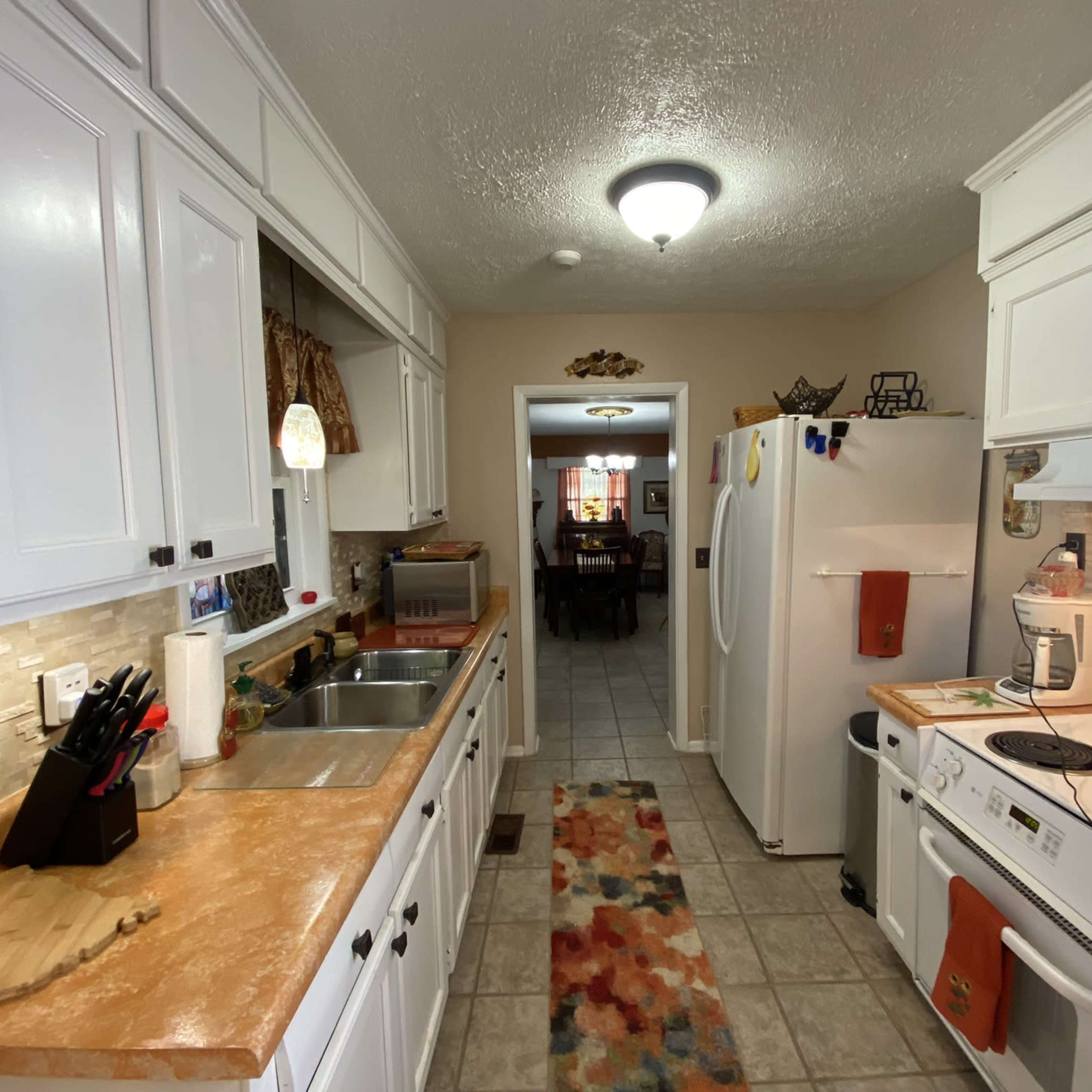 A compact kitchen with white cabinets, a dual sink, a stove, and a refrigerator, featuring tiled flooring and a colorful rug.