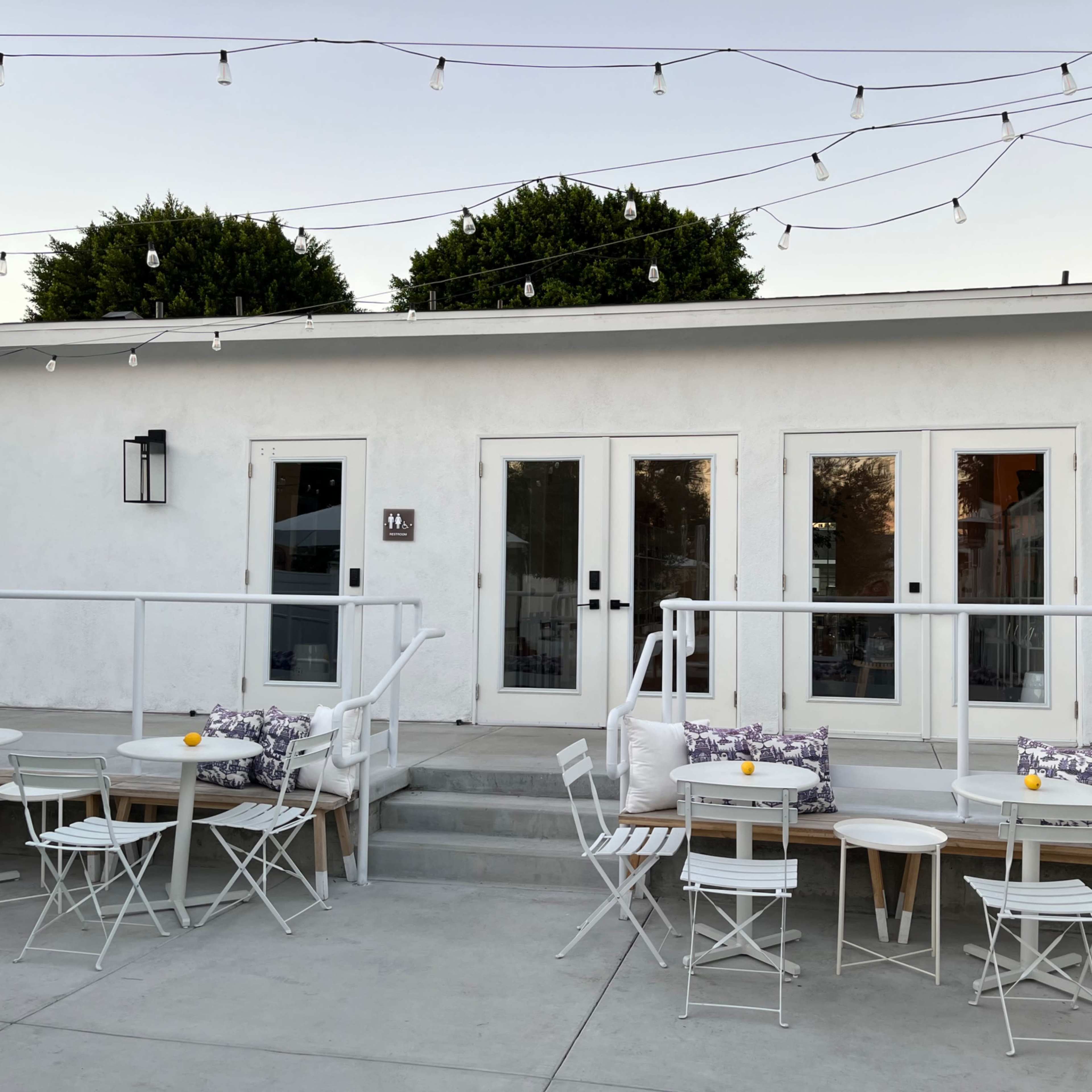 A modern outdoor patio area with white furniture and string lights illuminating a building with glass doors and a minimalist design.