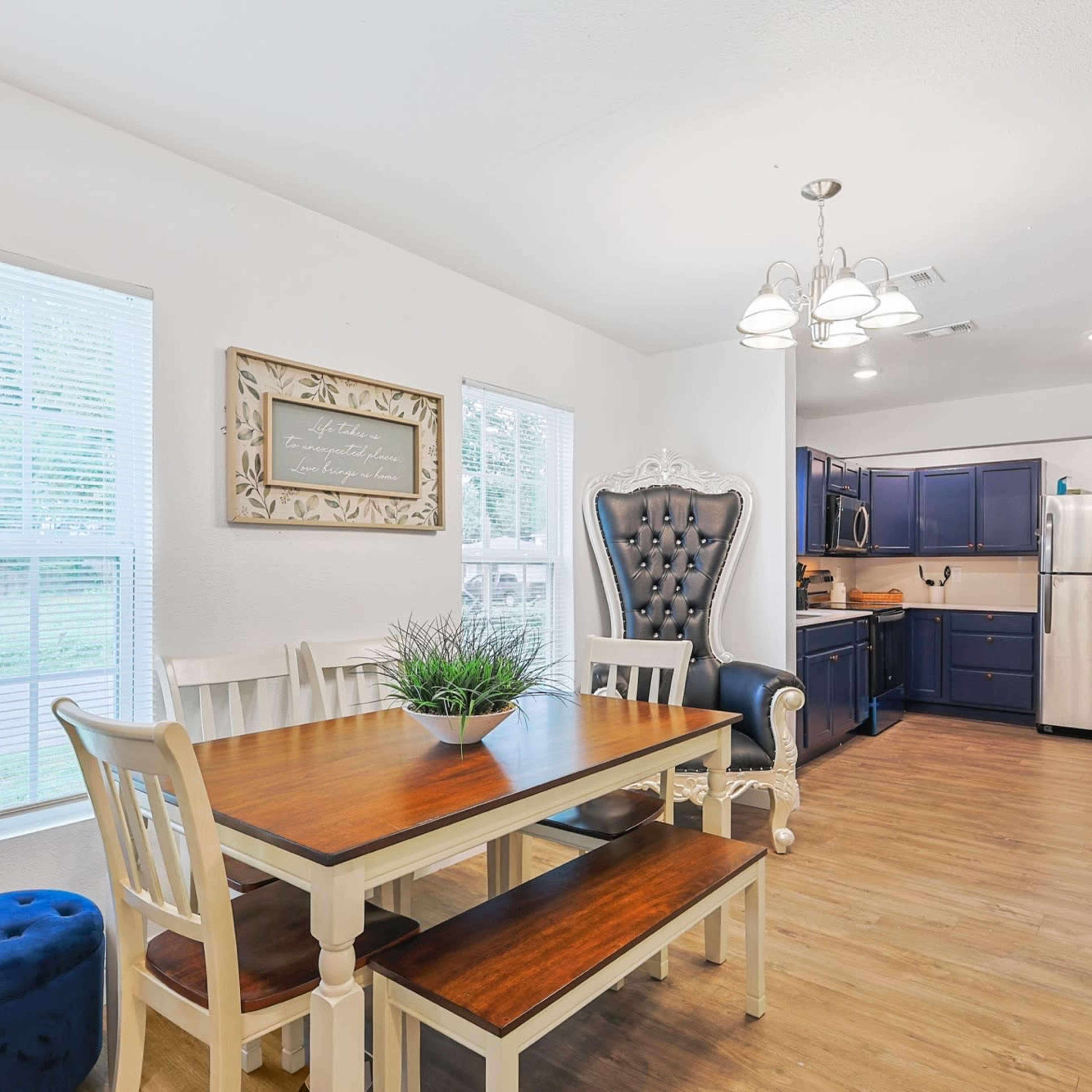 A dining area features a wooden table with seating, a large decorative chair, and a kitchen visible in the background.
