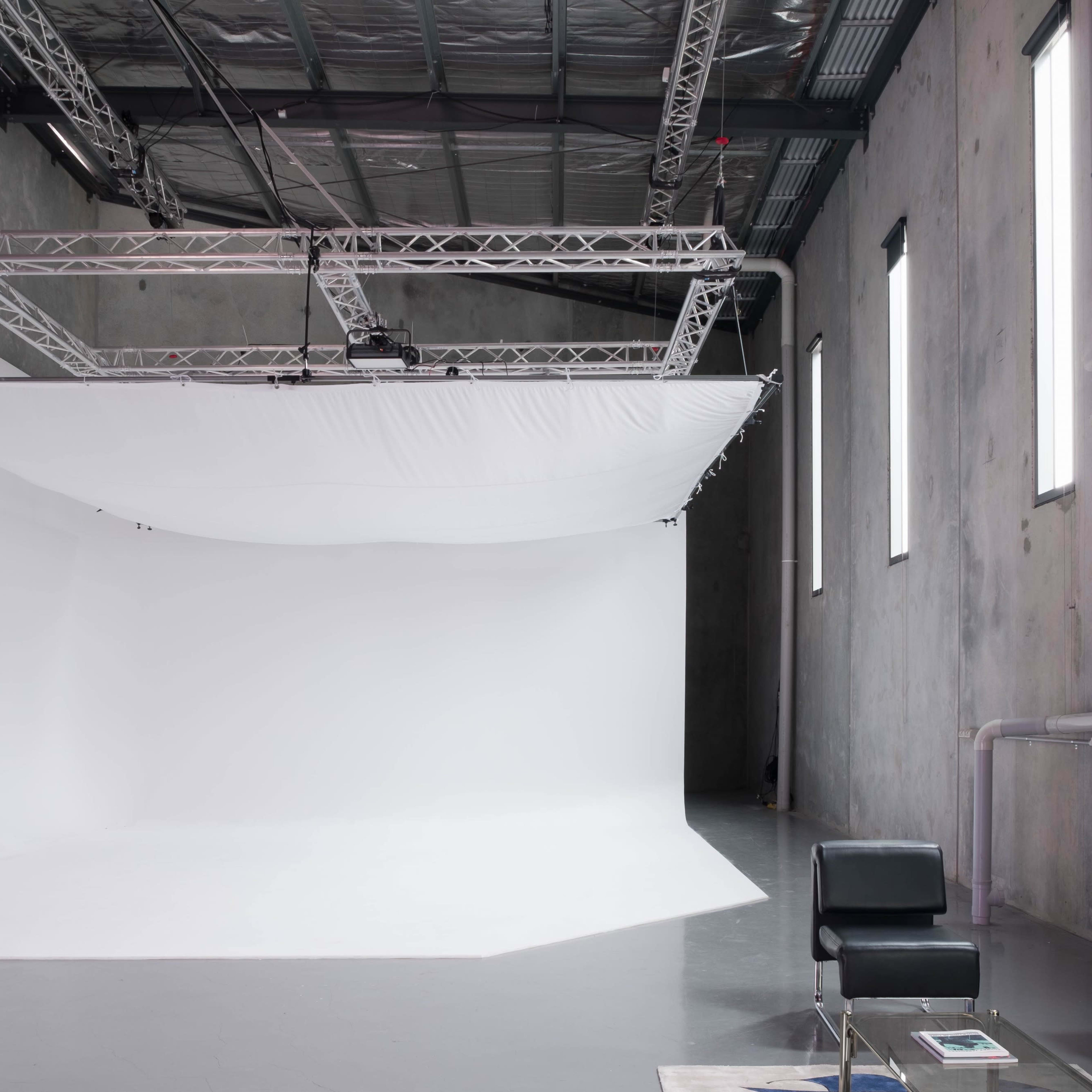 A photography studio with a large white backdrop, metal support structure overhead, and a black chair positioned near the edge.