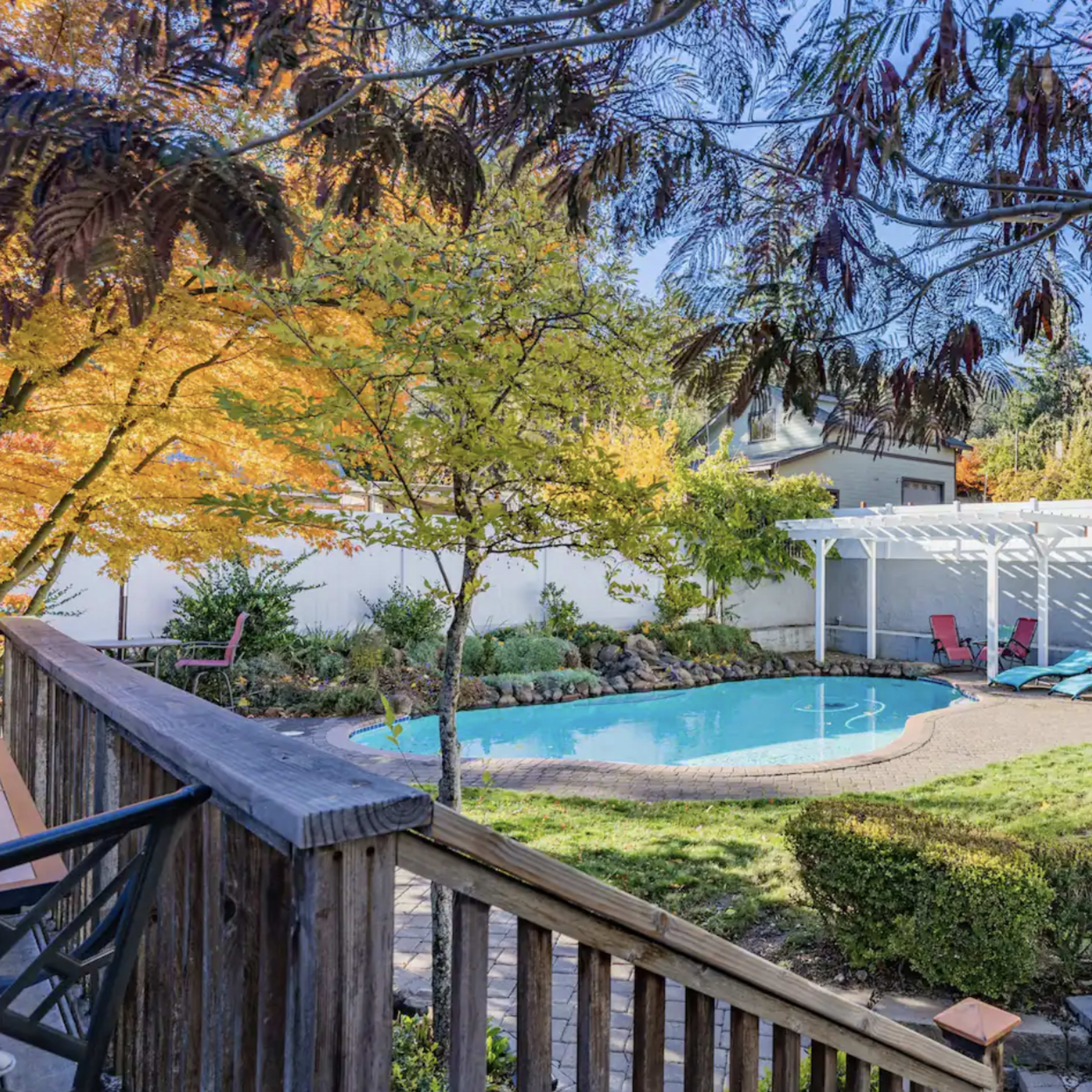 A backyard features a swimming pool surrounded by landscaped grass and a seating area, with colorful trees in the background.
