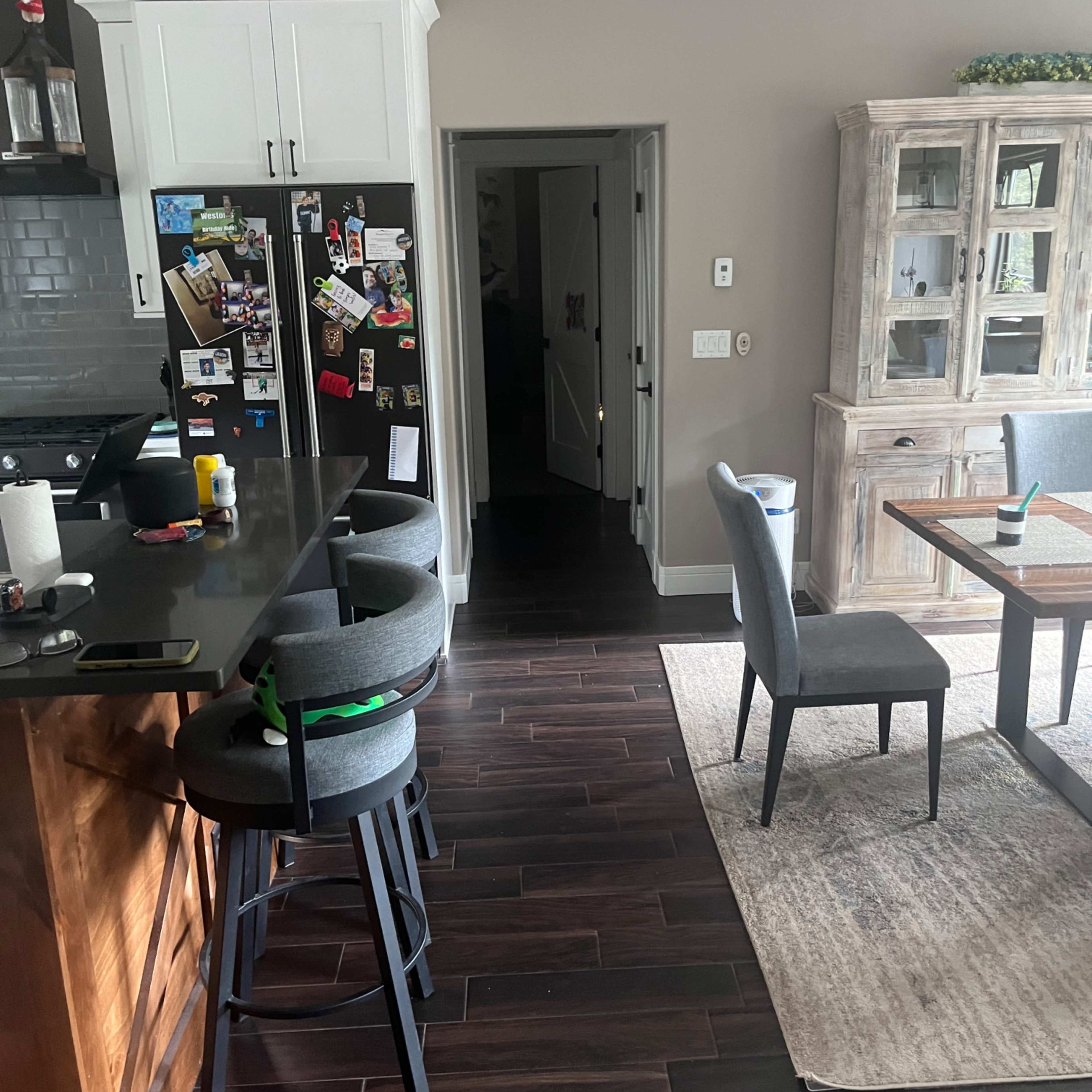 A modern kitchen with dark wooden flooring, a black countertop, a dining area with a table and chairs, and an open doorway leading to another room.