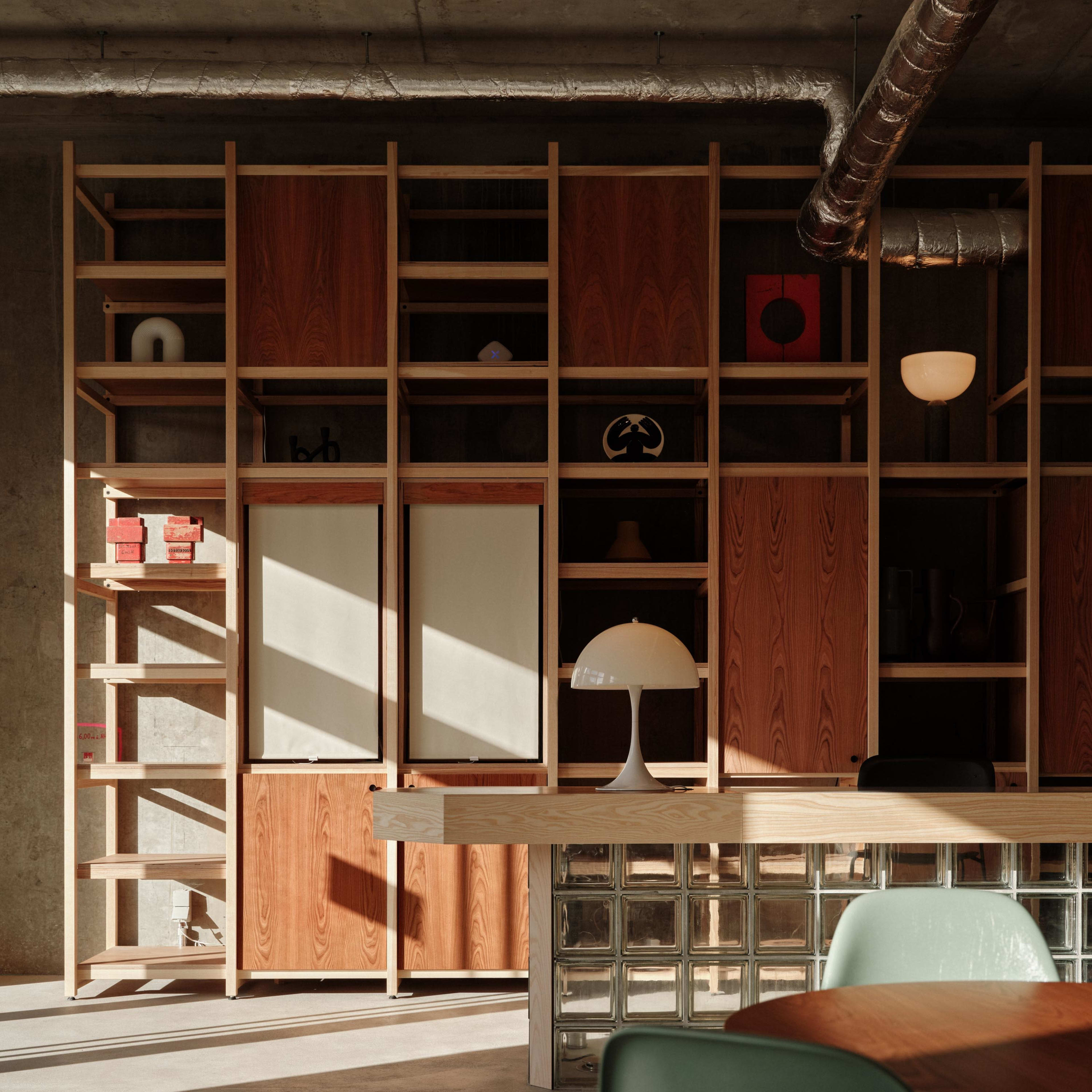 A modern interior with wooden shelving, a glass block wall, and a lamp on a table, illuminated by sunlight.