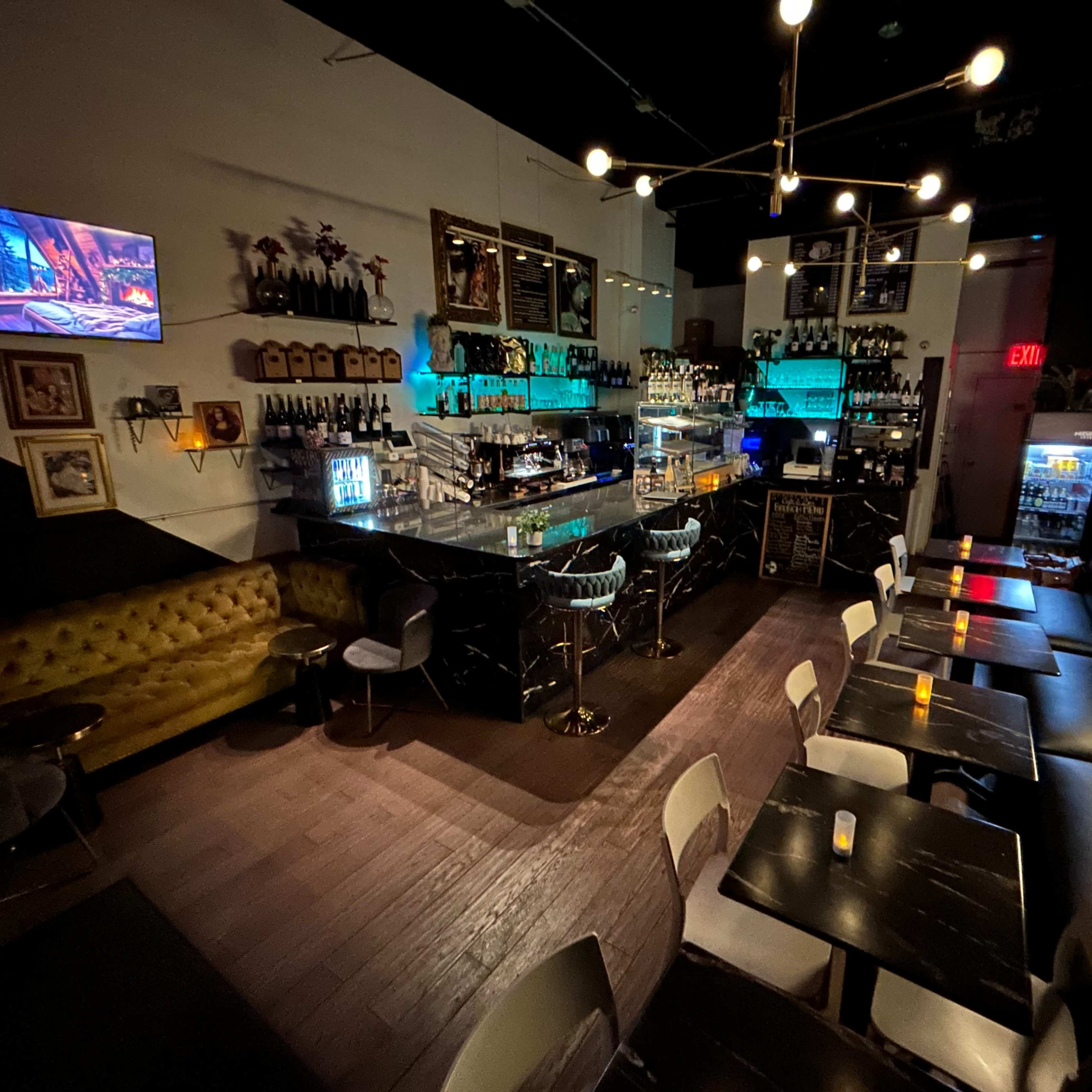 A dimly lit bar with a modern interior featuring a long countertop, rows of liquor bottles, and seating arrangements of sleek tables and chairs.