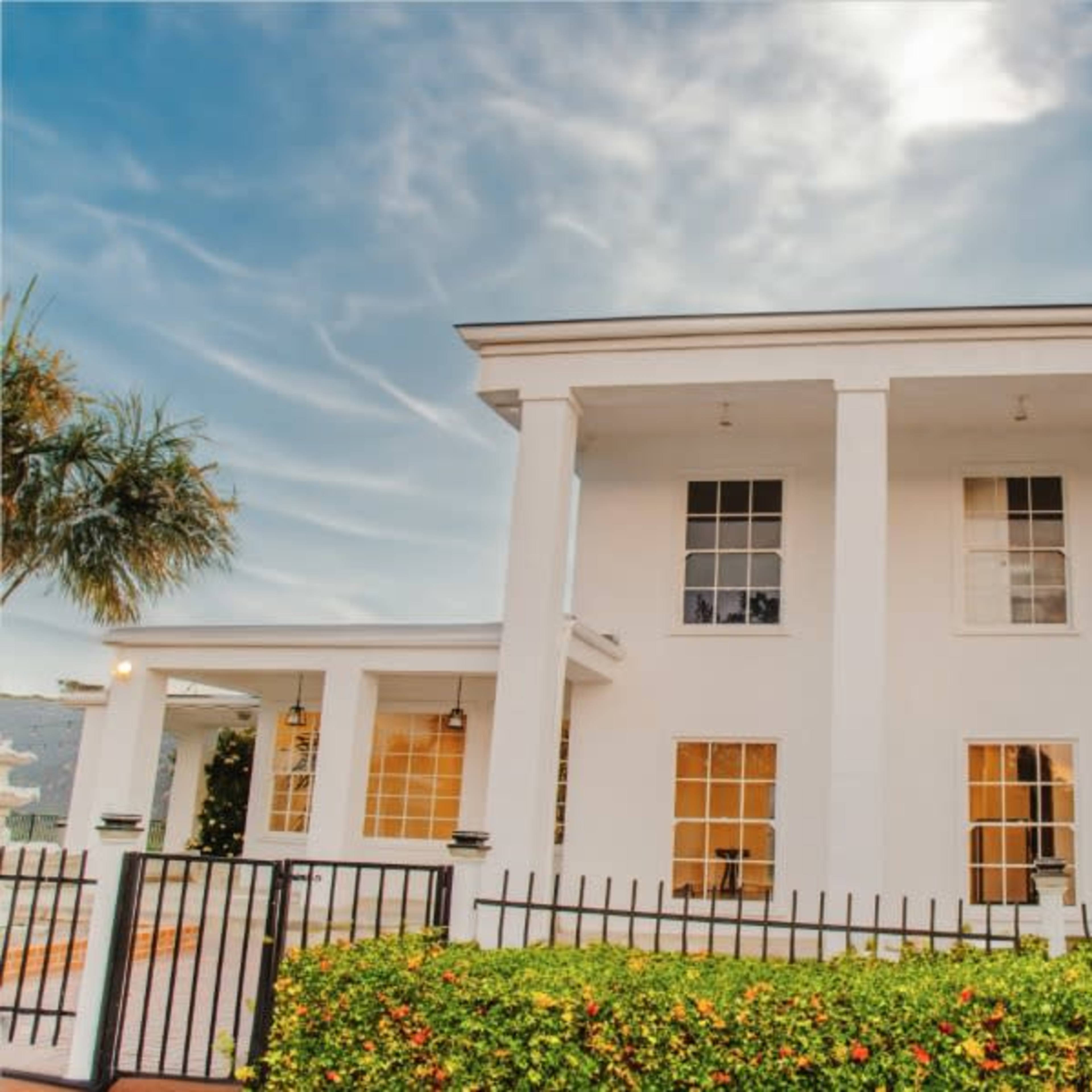 A two-story white house with tall columns and large windows, surrounded by a black metal fence and landscaped with green plants.